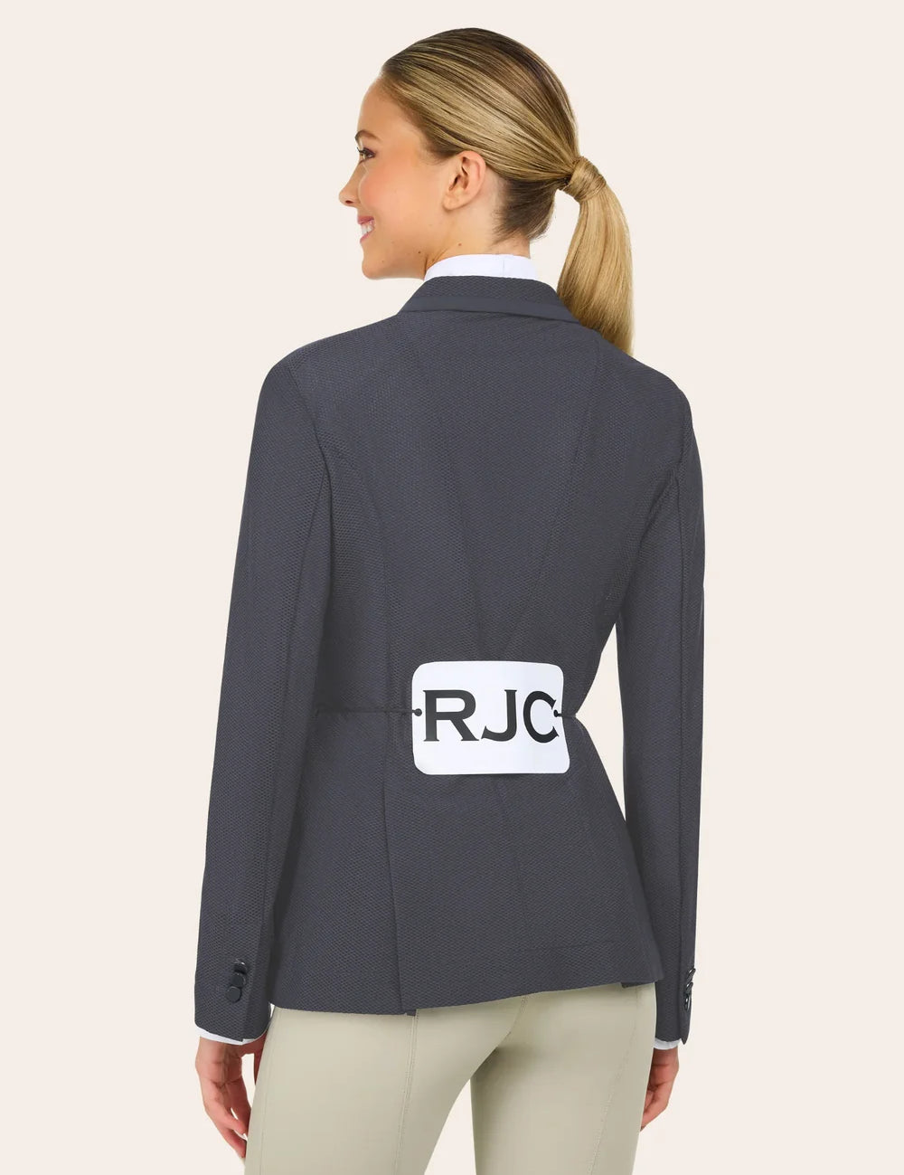 A woman with blond hair in a ponytail wears a dark gray blazer and tan pants, facing away, with a white label marked "RJC" pinned to her back.