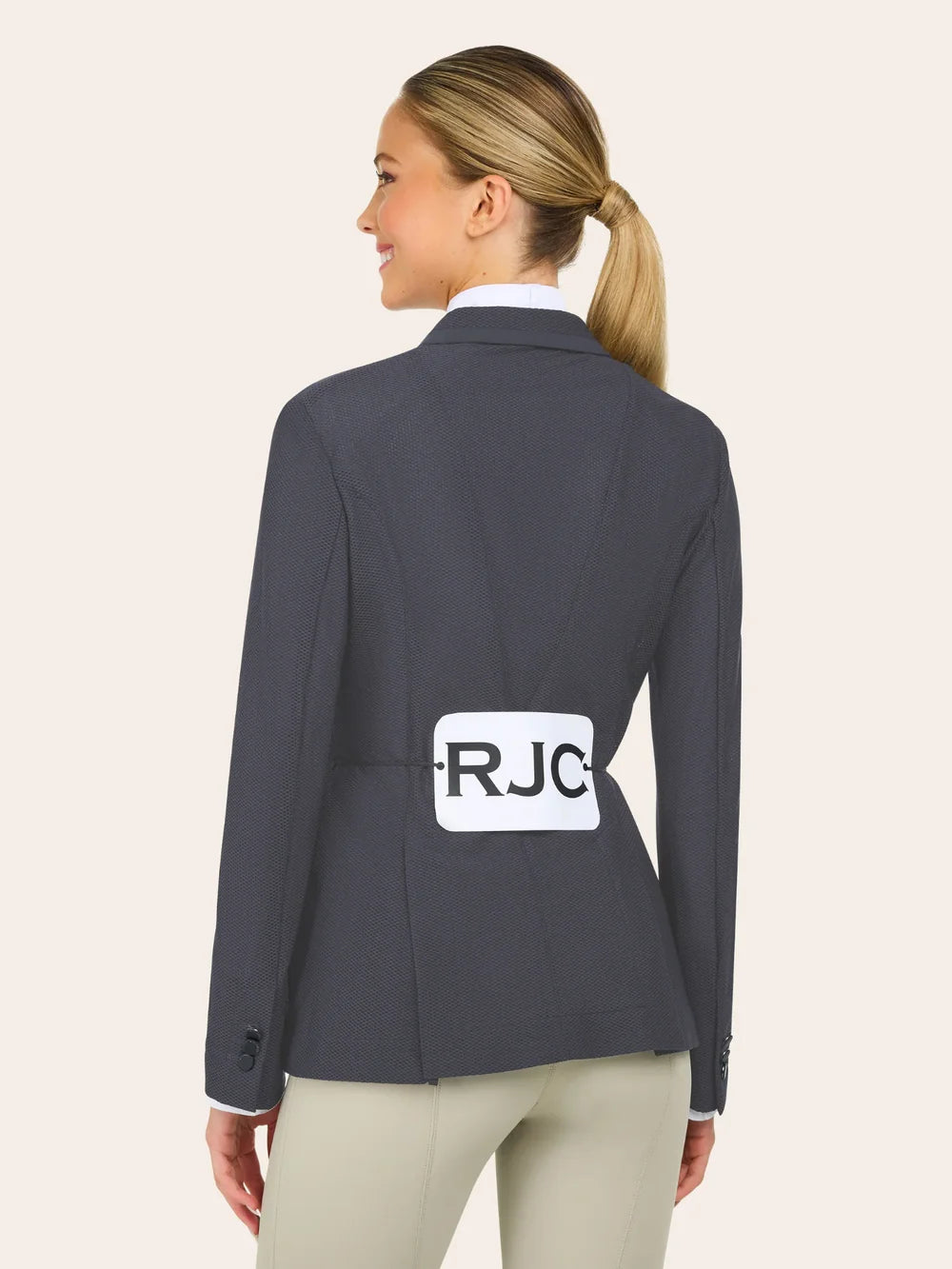 A woman with blond hair in a ponytail wears a dark gray blazer and tan pants, facing away, with a white label marked "RJC" pinned to her back.