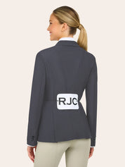 A woman with blond hair in a ponytail wears a dark gray blazer and tan pants, facing away, with a white label marked "RJC" pinned to her back.