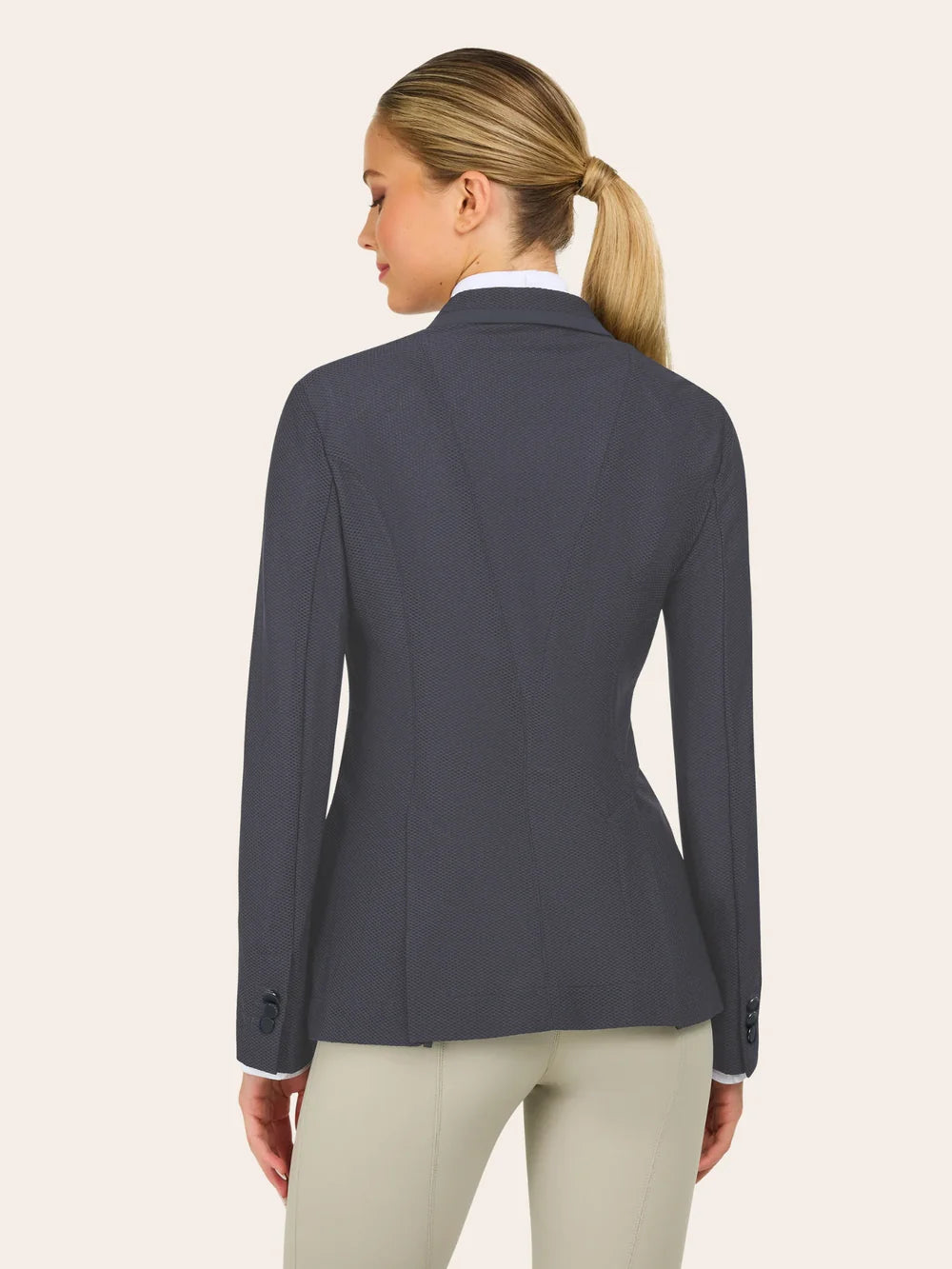Back view of a charcoal gray equestrian show jacket with a tailored, fitted silhouette, textured fabric, contoured seams, and buttoned cuffs, shown over a white show shirt with light tan riding breeches on a model against a plain light background.