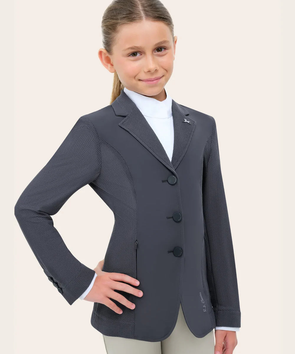 Angled front view of the RJ Classics Harmony Evo Jr. Mesh Show Coat in Odyssey Grey, a junior equestrian show jacket with breathable mesh sleeves and side panels, smooth front body, notched lapel with logo pin, and three-button closure, shown over a white show shirt with light tan riding breeches against a plain light background.