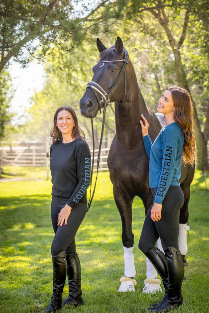 View of 2 female models wearing the Sailor Blue abd Black Kastel Crewneck Sweaters while standing outside in front of a black horse
