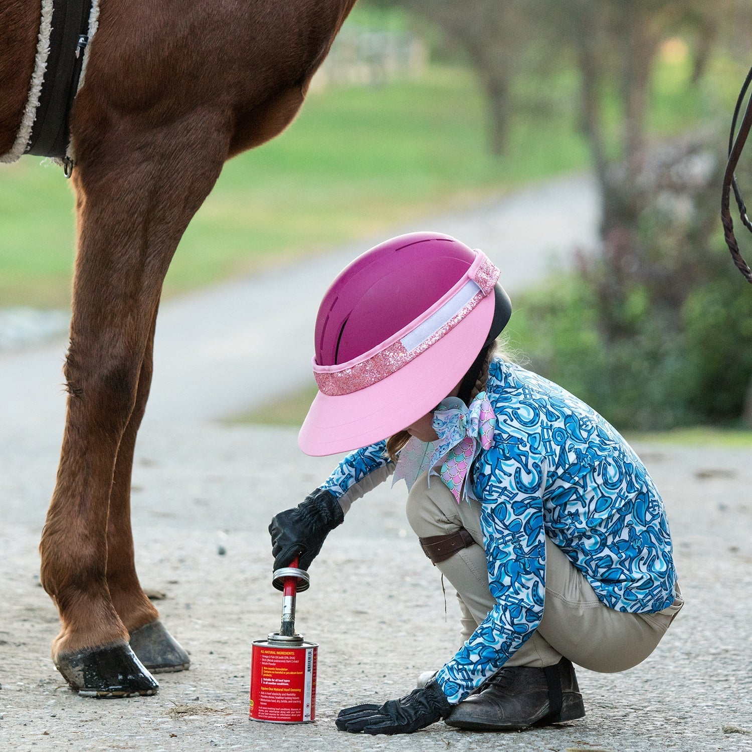 Side view of a young female model wearing the pink pizzazz EquiVisor Glitz Sun Visor as she's bending down to put hoof oil on a horse's hooves on a path outside