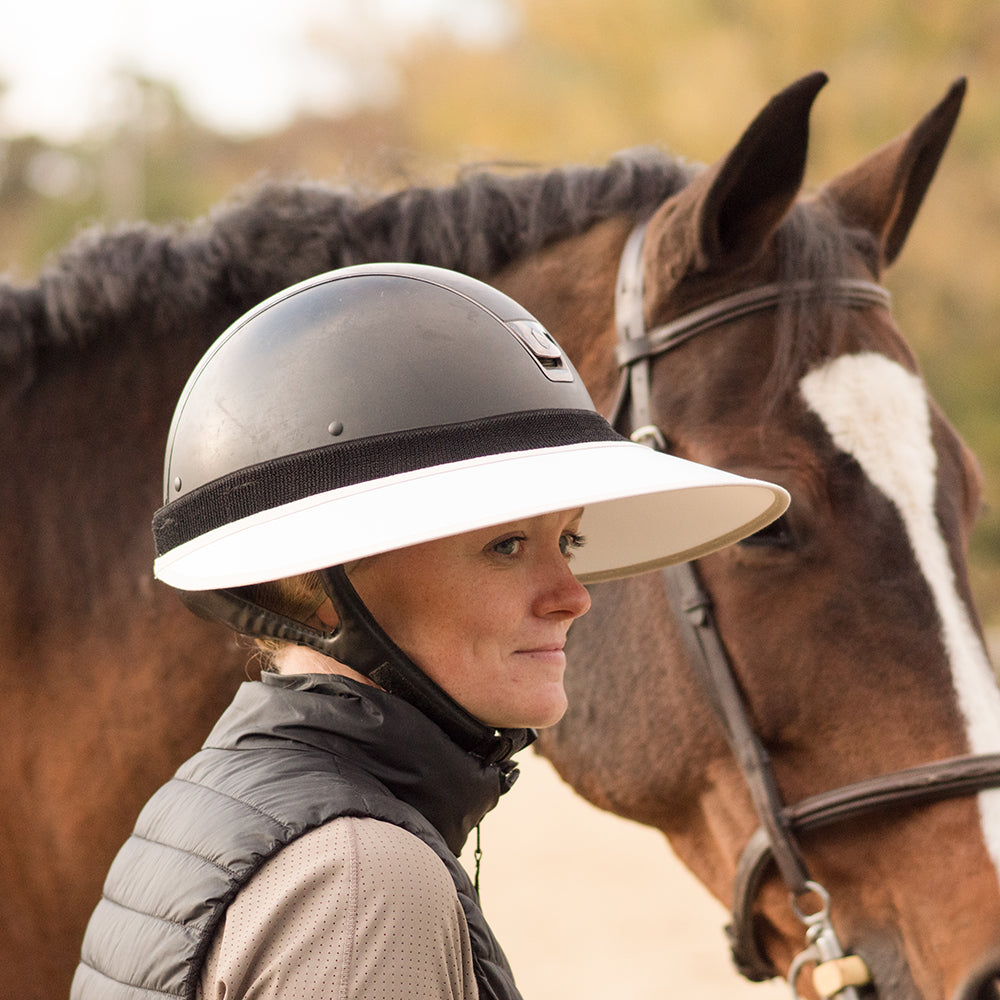 Side view of a female model wearing the white/mocha EquiVisor Sport Sun Visor while standing next to a horse