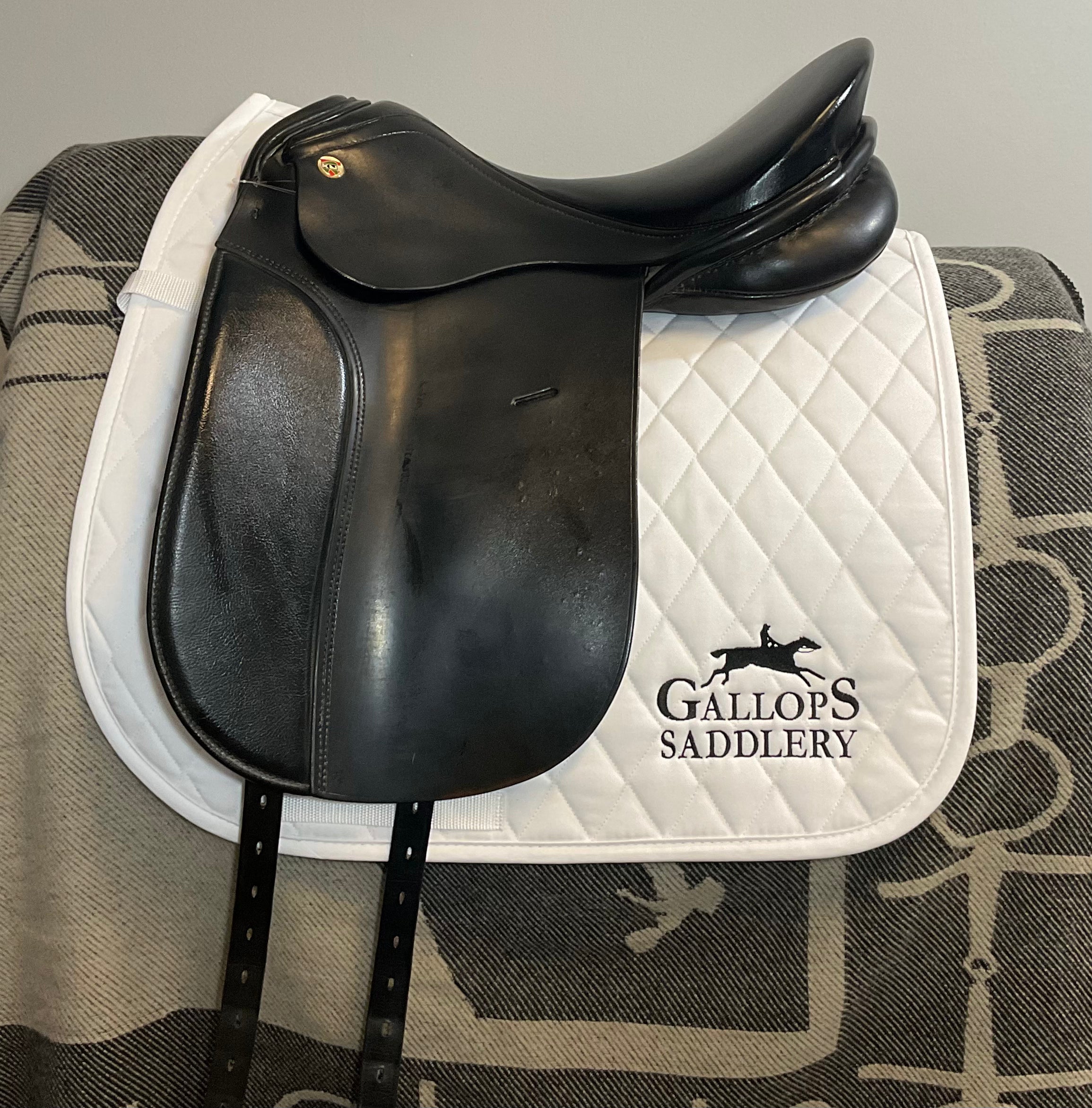 A black leather equestrian saddle resting on a white quilted saddle pad with the 'GALLOP'S SADDLERY' logo printed in black, set against a gray patterned blanket.