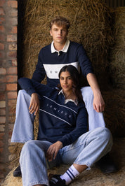 Two models wearing navy LeMieux rugby shirts with white collar and chest panel, seated on hay bales in a barn setting.