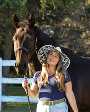 Front view of a female model wearing the Dreamers & Schemers Hay Sunshine F Bomb Hat while standing in front of a black horse