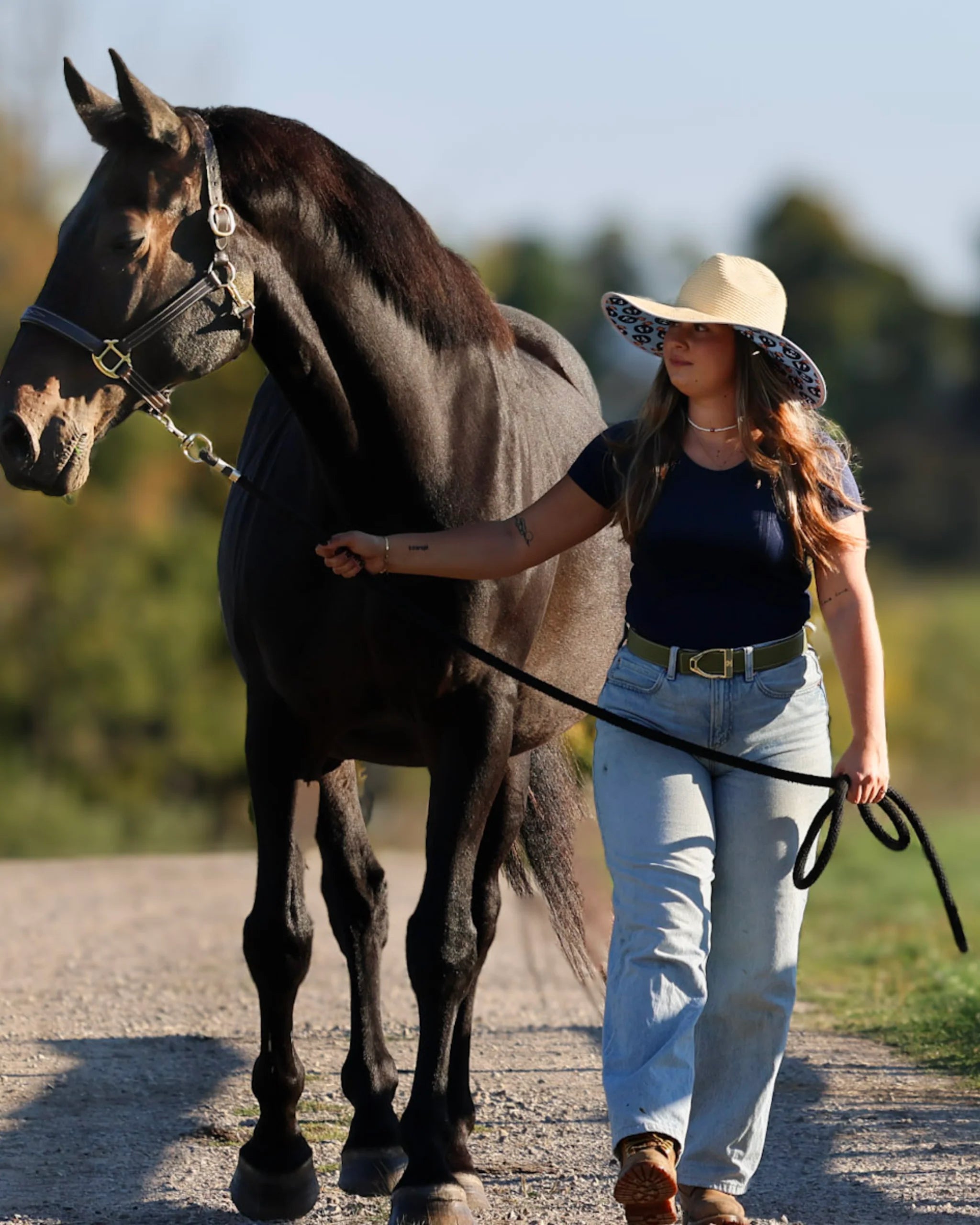 Front view of a female model wearing the Dreamers & Schemers Hay Sunshine F Bomb Hat while leading a black horse on a path outside