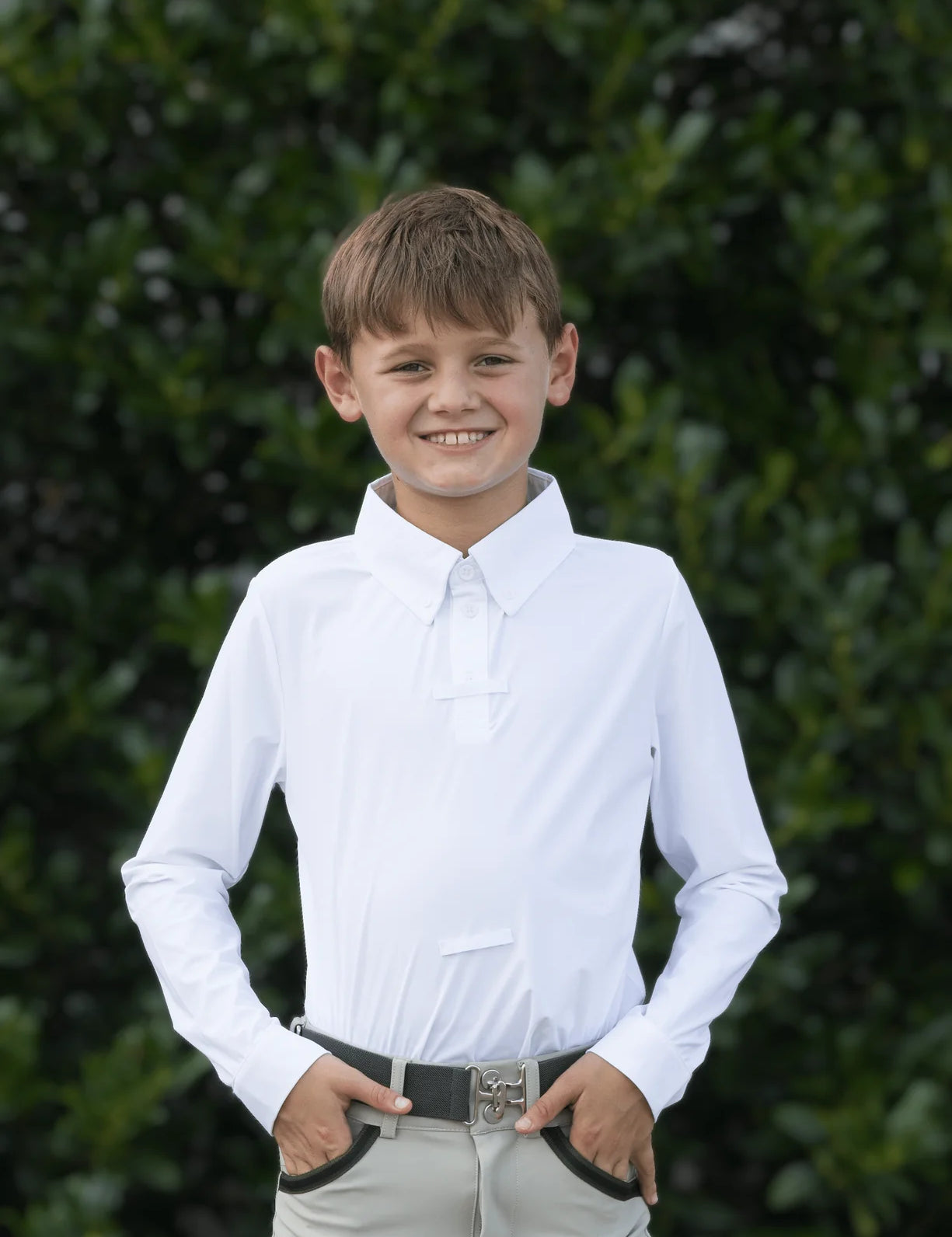 Front view of a young male model wearing the white Free Ride Equestrian Boy's Competition Shirt with green bushes in the background