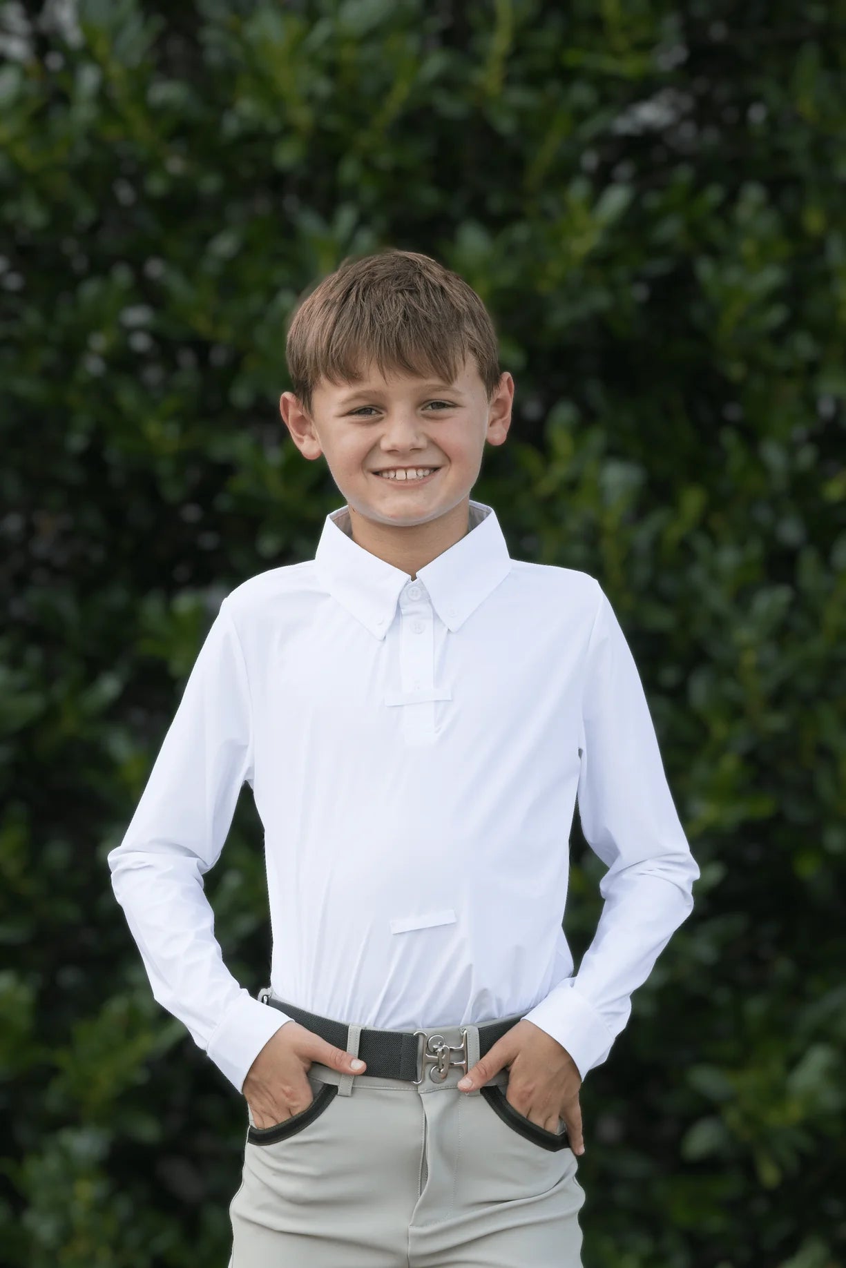 Front view of a young male model wearing the white Free Ride Equestrian Boy's Competition Shirt with green bushes in the background