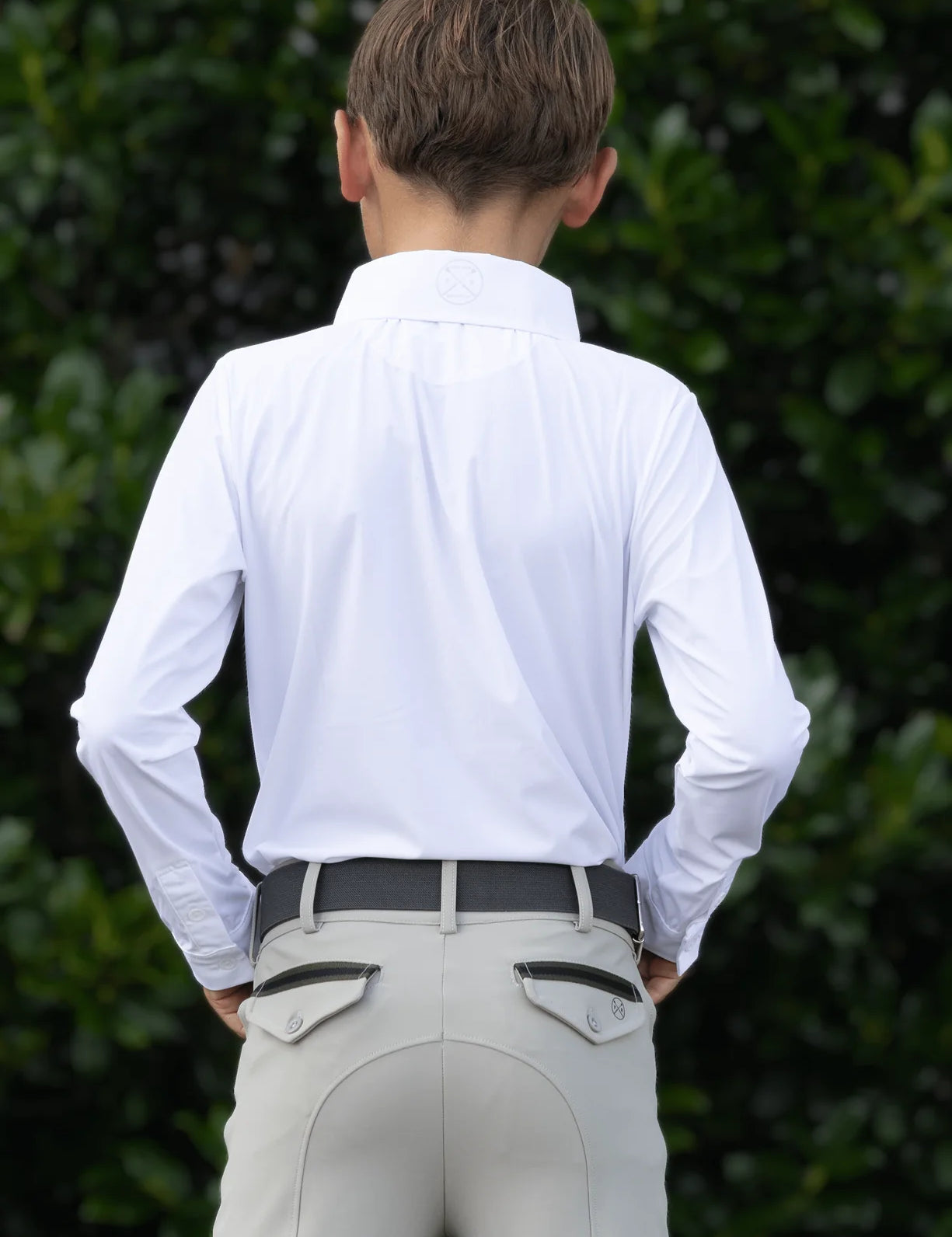 Back view of a young male model wearing the white Free Ride Equestrian Boy's Competition Shirt with green bushes in the background