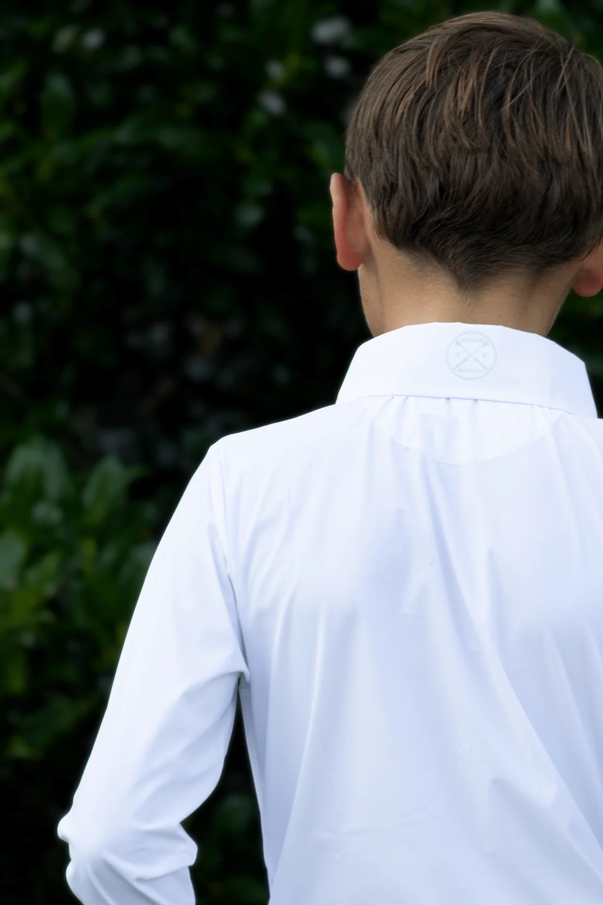 Close-up back view of a young male model wearing the white Free Ride Equestrian Boy's Competition Shirt with green bushes in the background