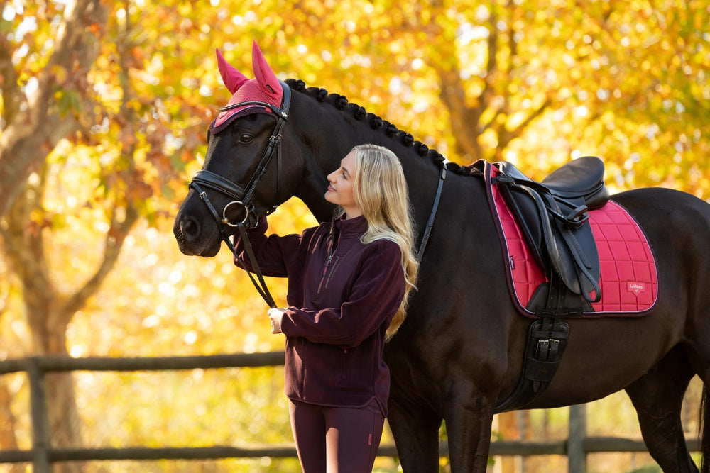 Side view of a female model standing in front of a dark colored horse wearing the cranberry LeMieux Classic Fly Hood while standing outside