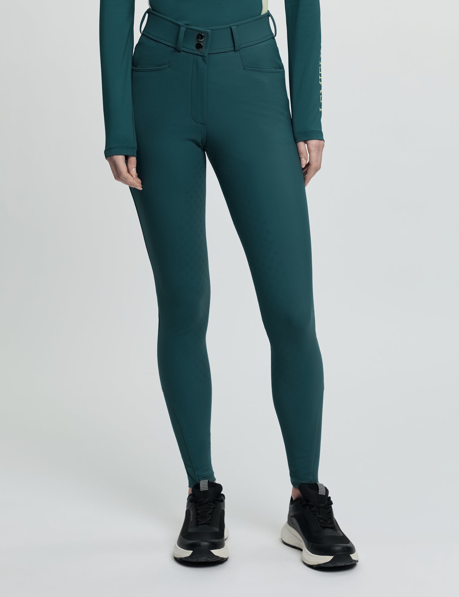 Model wearing LeMieux Isabelle Full Seat Breeches in green with high-rise waistband and black athletic shoes