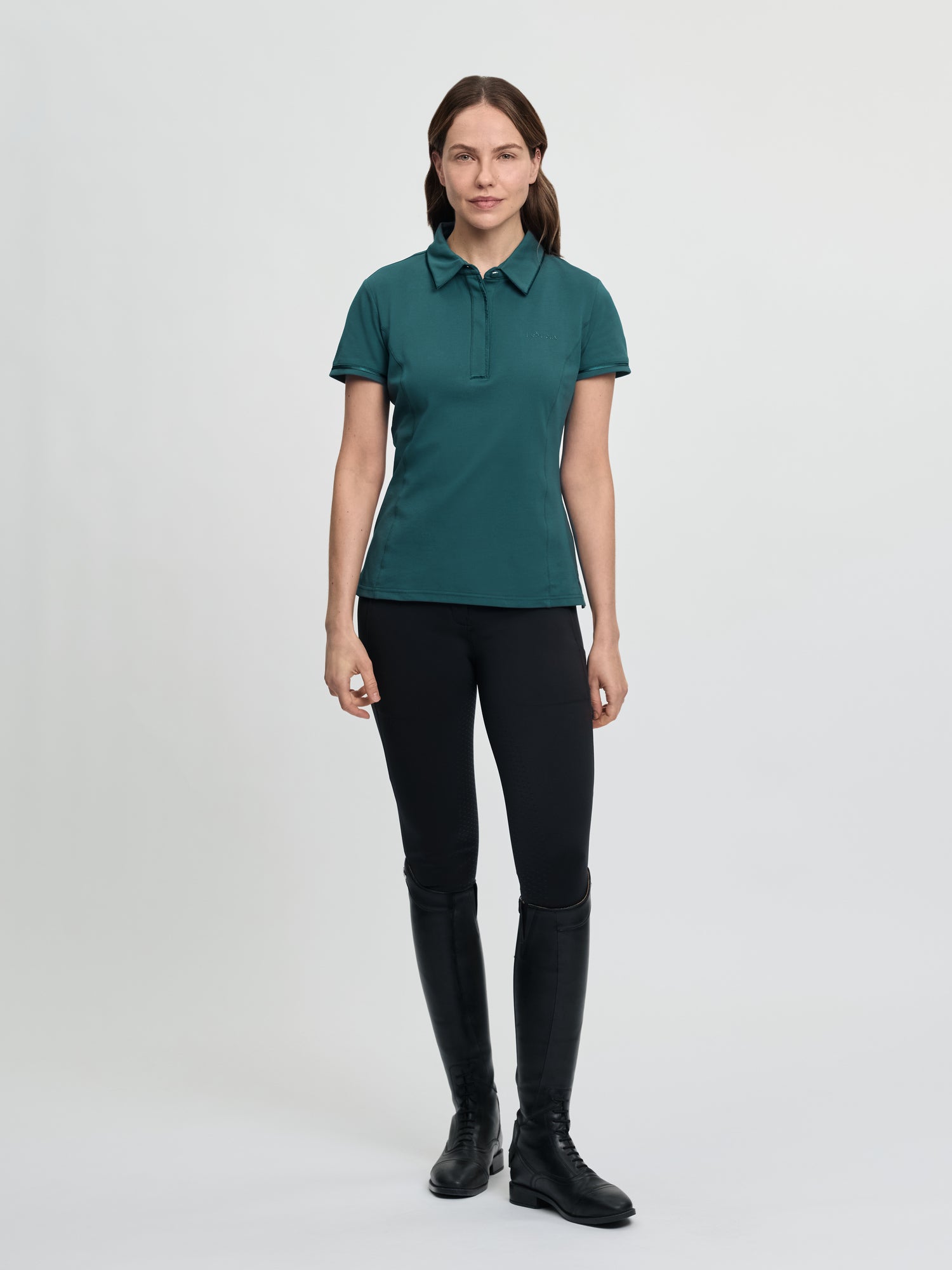 Full-body view of the LeMieux Pixie polo shirt in jungle green with a pointed collar, partial button placket, and short sleeves, worn with black tights and tall black riding boots against a light gray background.