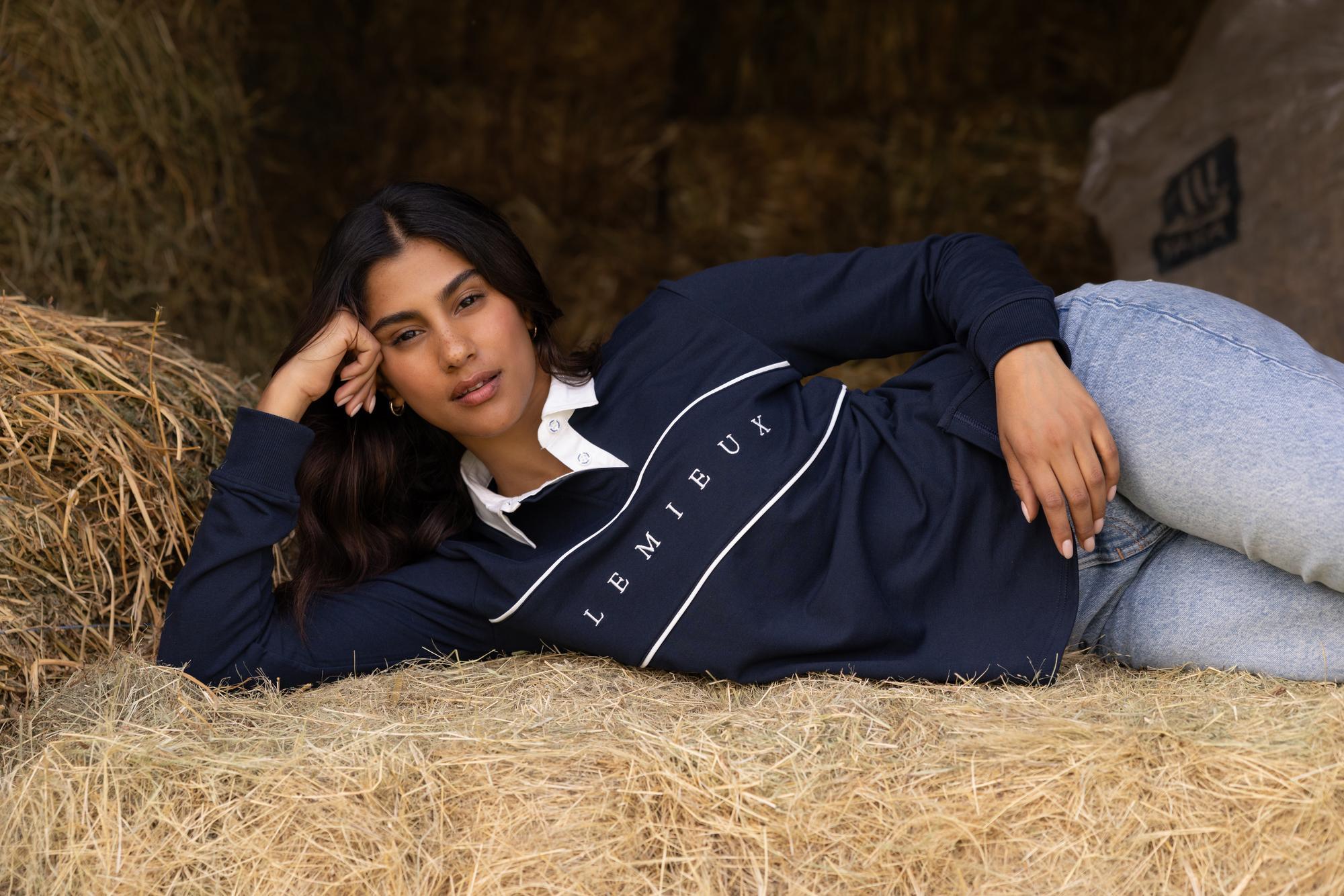 Model lying on hay wearing navy LeMieux Rochelle Rugby Shirt with white collar and chest piping detail.