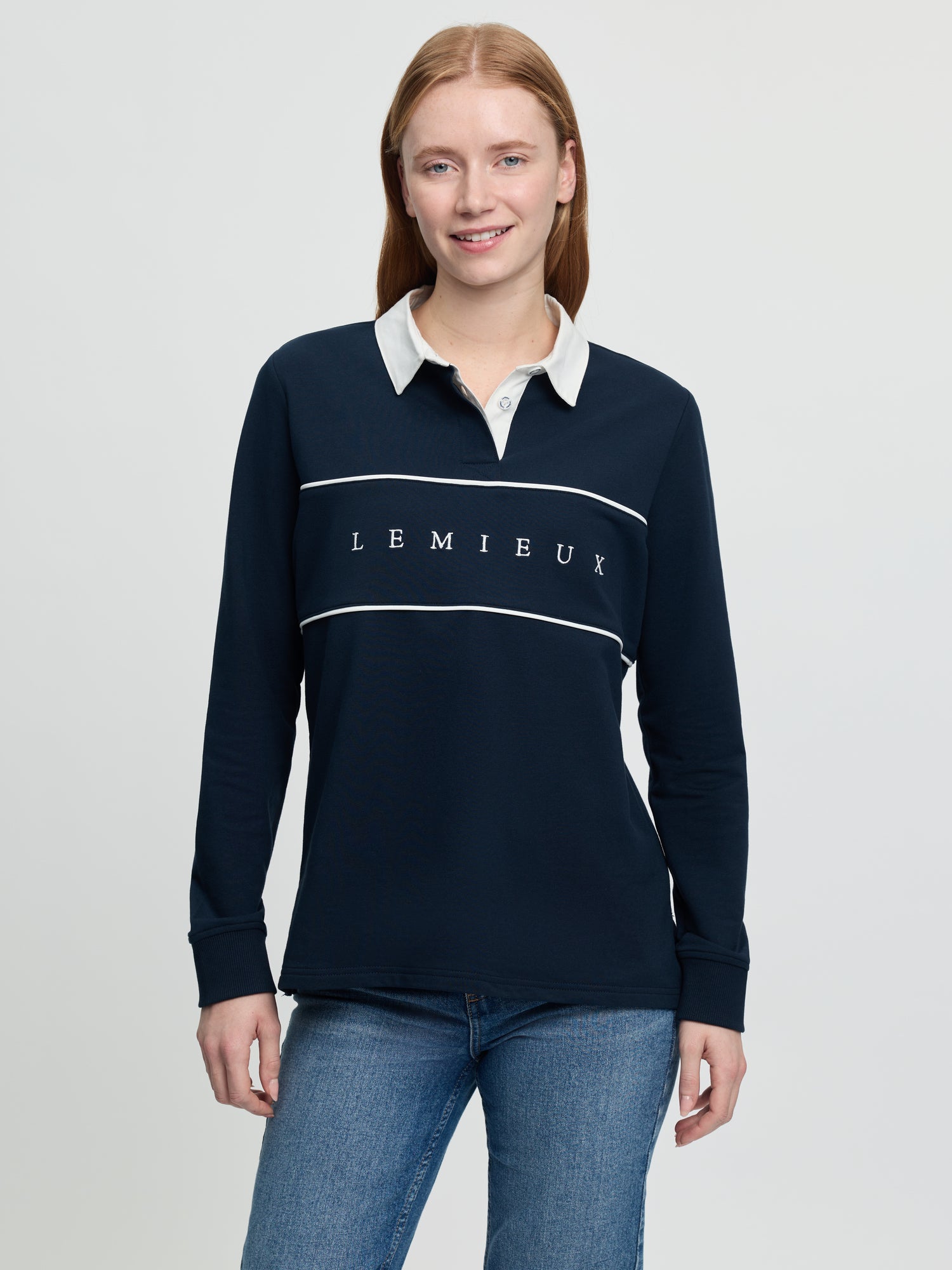 Front view of the LeMieux Rochelle rugby shirt in navy featuring a white collar, long sleeves with ribbed cuffs, white piping across the chest, and “LEMIEUX” lettering centered on the front, worn with blue jeans against a light gray background.