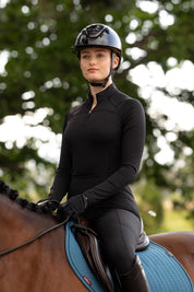 Outdoor photo of the LeMieux Airflow Long Sleeve Base Layer in black, showing a quarter-zip high collar and long sleeves, worn while riding a horse with a black helmet and gloves.