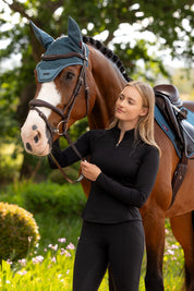 LeMieux Airflow long sleeve base layer in black with quarter-zip high collar, worn with black riding tights while standing beside a saddled horse outdoors.