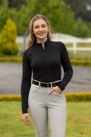 Outdoor photo of the LeMieux Airflow Long Sleeve Base Layer in black, showing a quarter-zip high collar and long sleeves, worn with light gray breeches and a black belt.