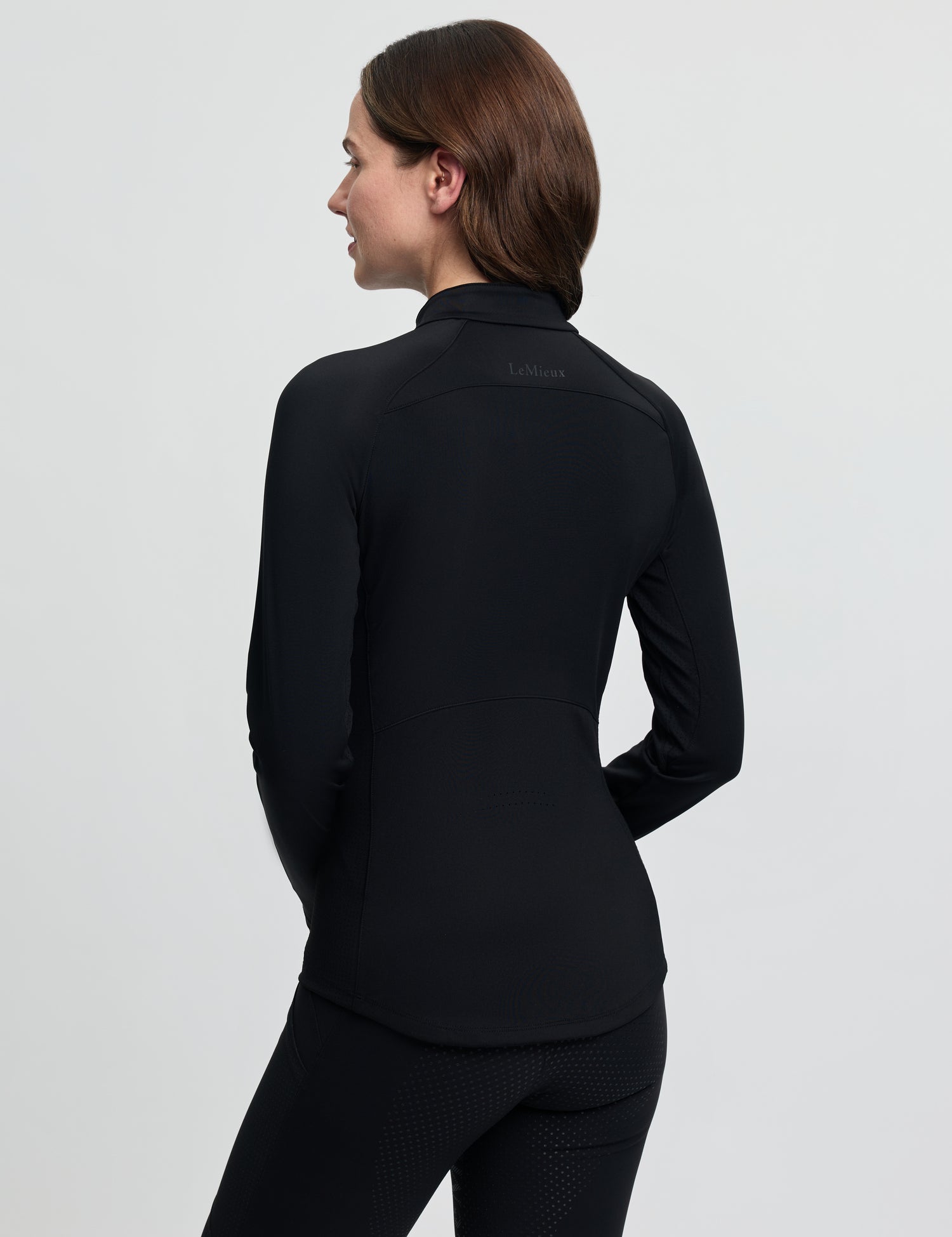Back view of the LeMieux Airflow Long Sleeve Base Layer in black, showing a fitted silhouette with a high collar and seam detailing, worn with black riding tights against a light gray background.