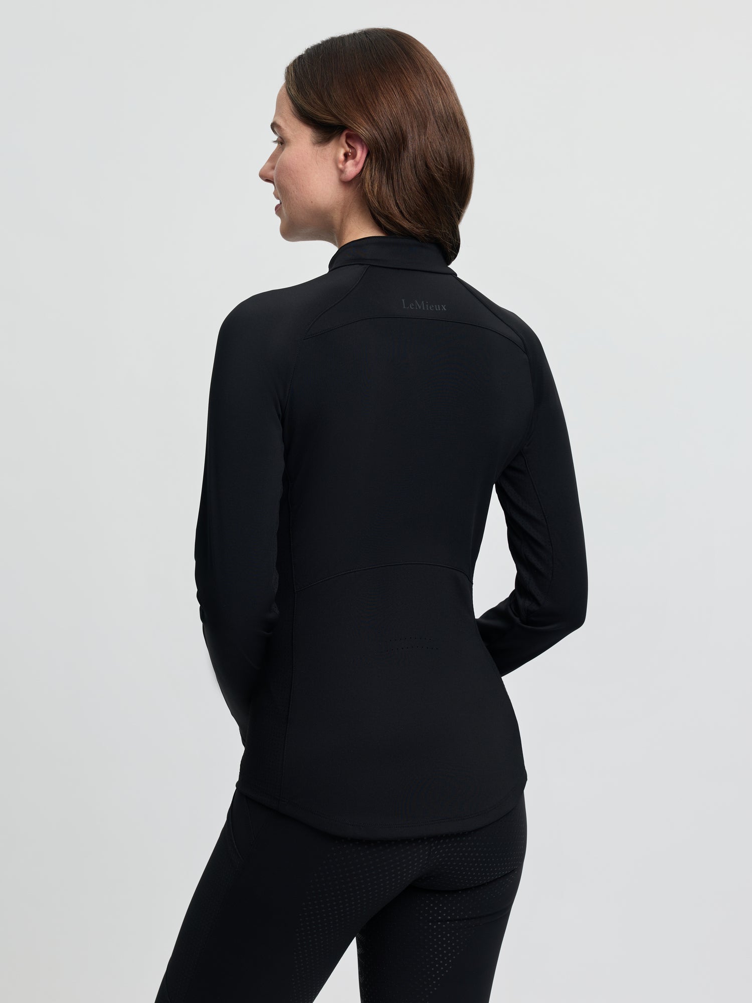 Back view of the LeMieux Airflow Long Sleeve Base Layer in black, showing a fitted silhouette with a high collar and seam detailing, worn with black riding tights against a light gray background.