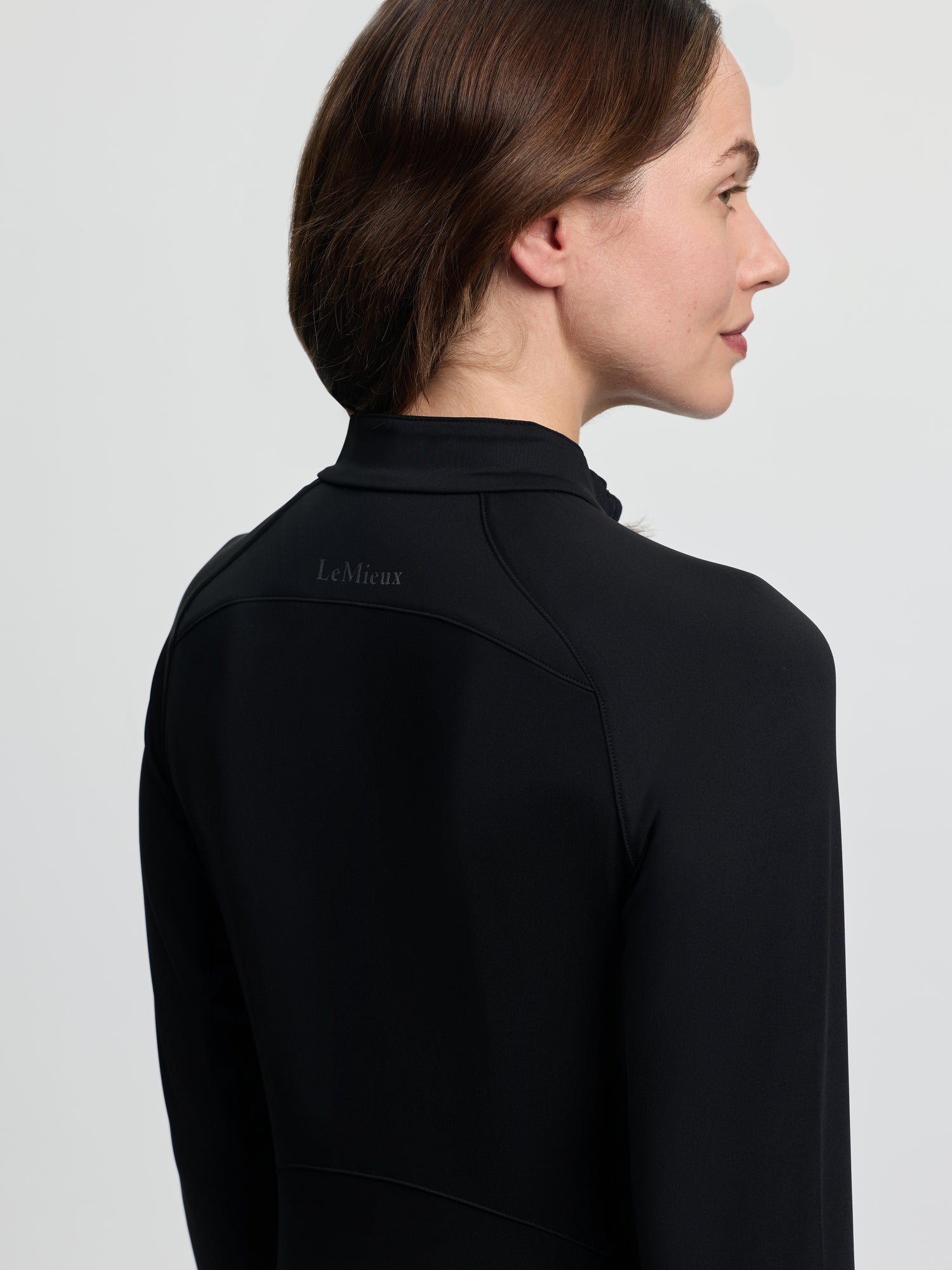 Close-up back/side view of the LeMieux Airflow Long Sleeve Base Layer in black, showing the high collar, shoulder seam lines, and subtle LeMieux logo on the upper back against a light gray background.
