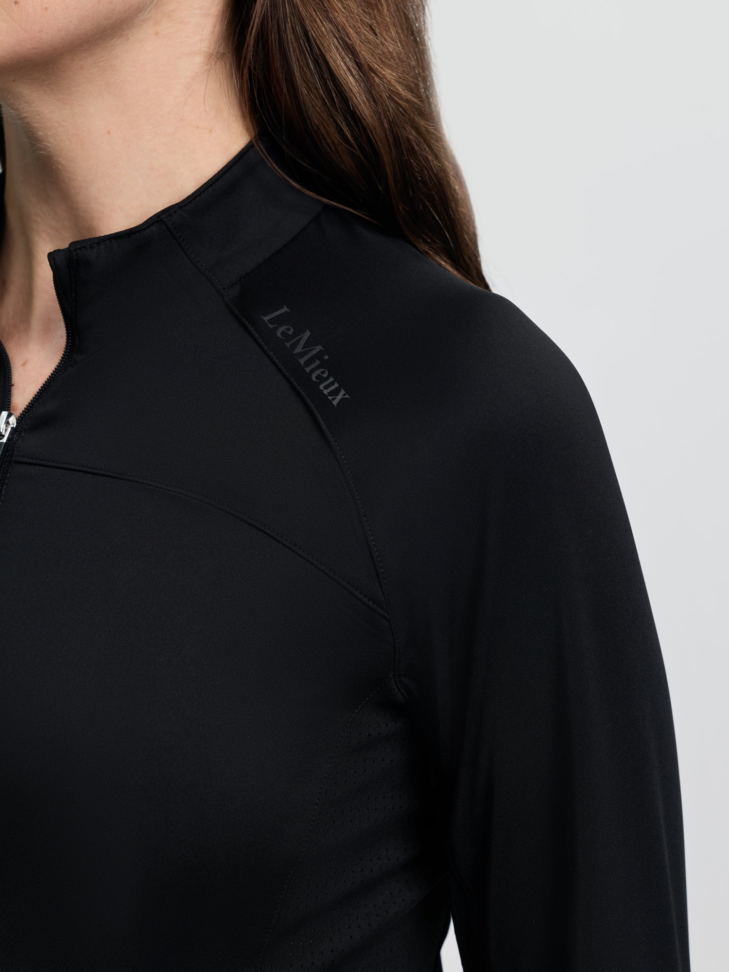 Close-up of the LeMieux Airflow Long Sleeve Base Layer in black showing the quarter-zip high collar, seam detailing, and LeMieux logo on the shoulder against a light gray background.