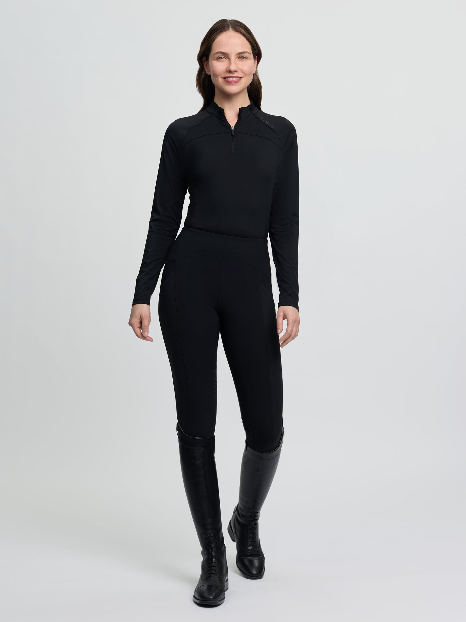 Full-body view of the LeMieux Airflow Long Sleeve Base Layer in black, featuring a high collar with quarter-zip front and long sleeves, shown on a model wearing black riding tights and tall black riding boots against a light gray background.