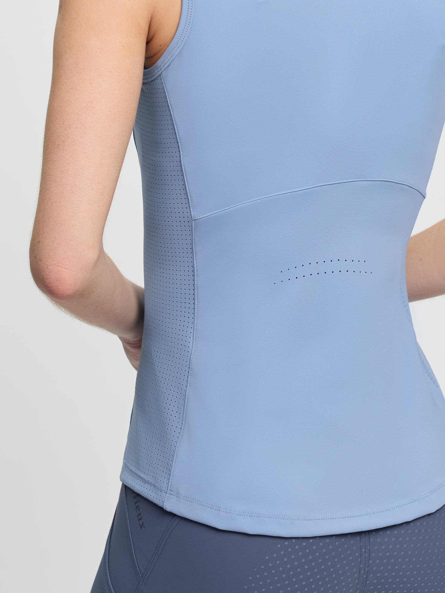 Close-up back view of the LeMieux Airflow sleeveless base layer in powder blue showing a perforated panel at mid-back and a textured side panel, worn with blue riding tights against a light gray background.