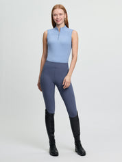 Full-body view of the LeMieux Airflow sleeveless base layer in powder blue with a quarter-zip high collar, worn with blue riding tights and tall black riding boots against a light gray background.