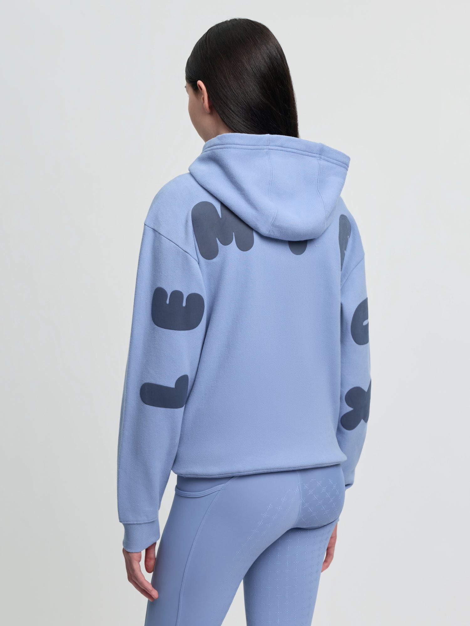 Angled back view of model wearing LeMieux Young Rider Sia Hoodie in powder blue showing hood and oversized sleeve lettering