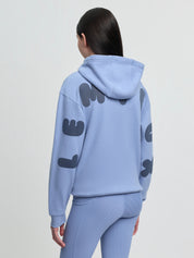 Angled back view of model wearing LeMieux Young Rider Sia Hoodie in powder blue showing hood and oversized sleeve lettering