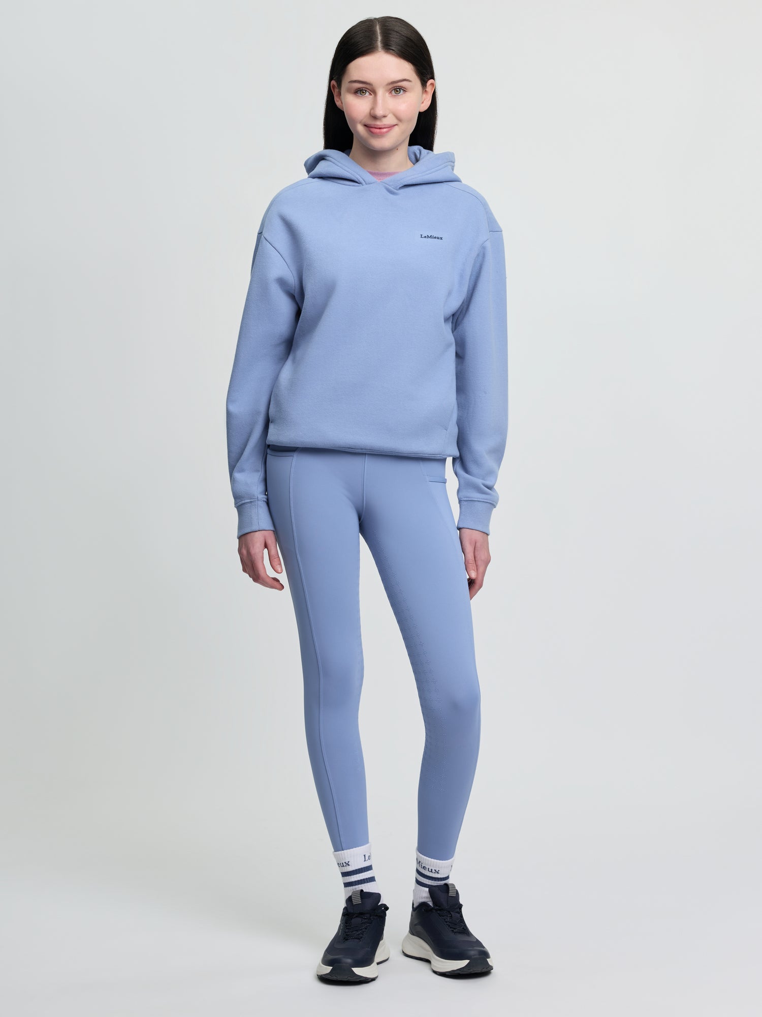 Model wearing LeMieux Young Rider Sia Hoodie in powder blue with matching leggings, striped socks, and black athletic shoes