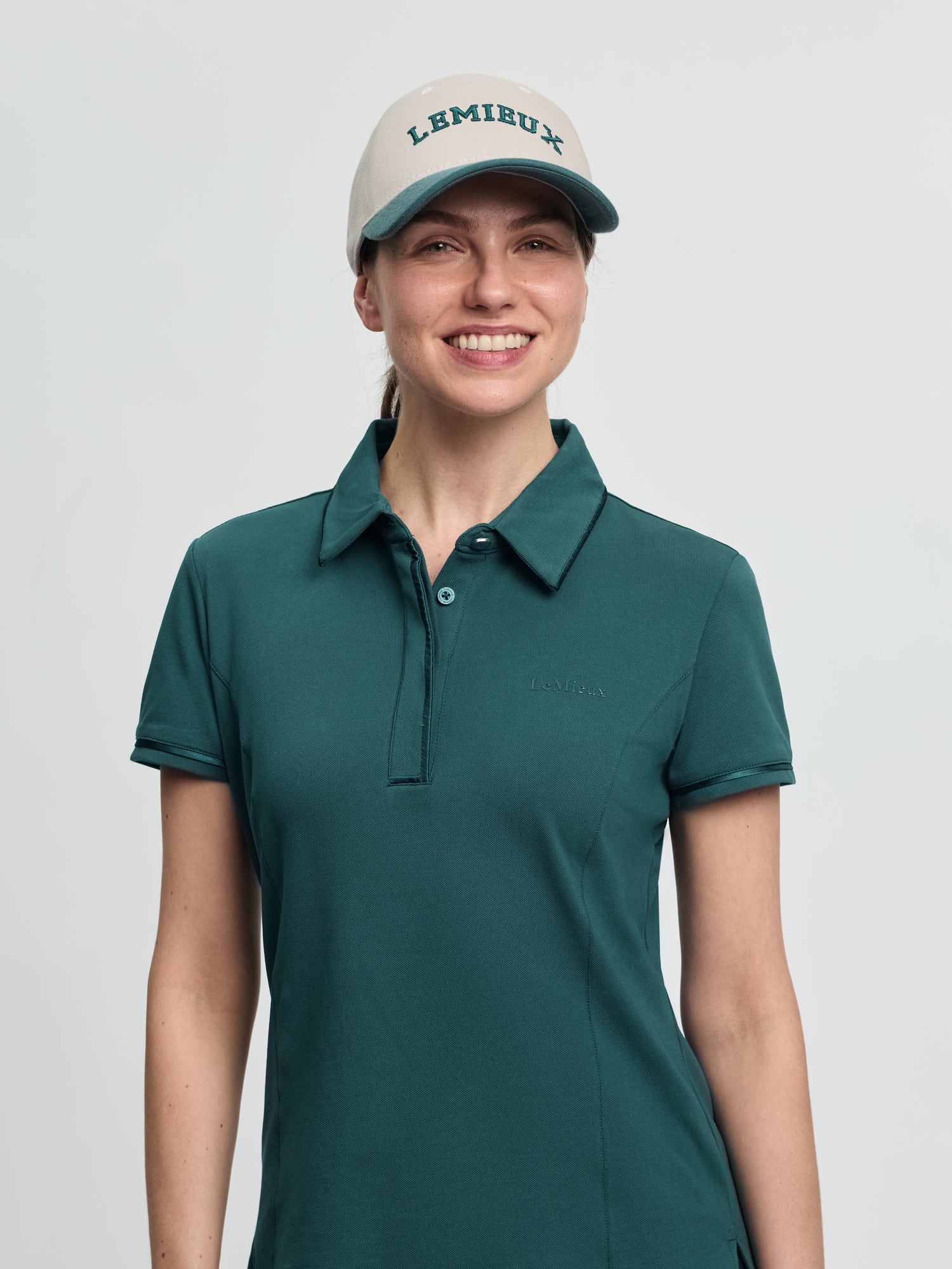 LeMieux Pixie polo shirt in jungle green with a pointed collar, partial button placket, and short sleeves, worn with a light cap with “LEMIEUX” lettering against a light gray background.