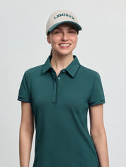 LeMieux Pixie polo shirt in jungle green with a pointed collar, partial button placket, and short sleeves, worn with a light cap with “LEMIEUX” lettering against a light gray background.
