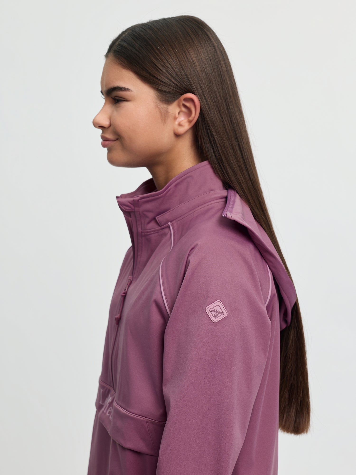 Side view of model wearing LeMieux Young Rider Paige Quarter Zip Waterproof Jacket in Mallow showing hood and sleeve logo patch