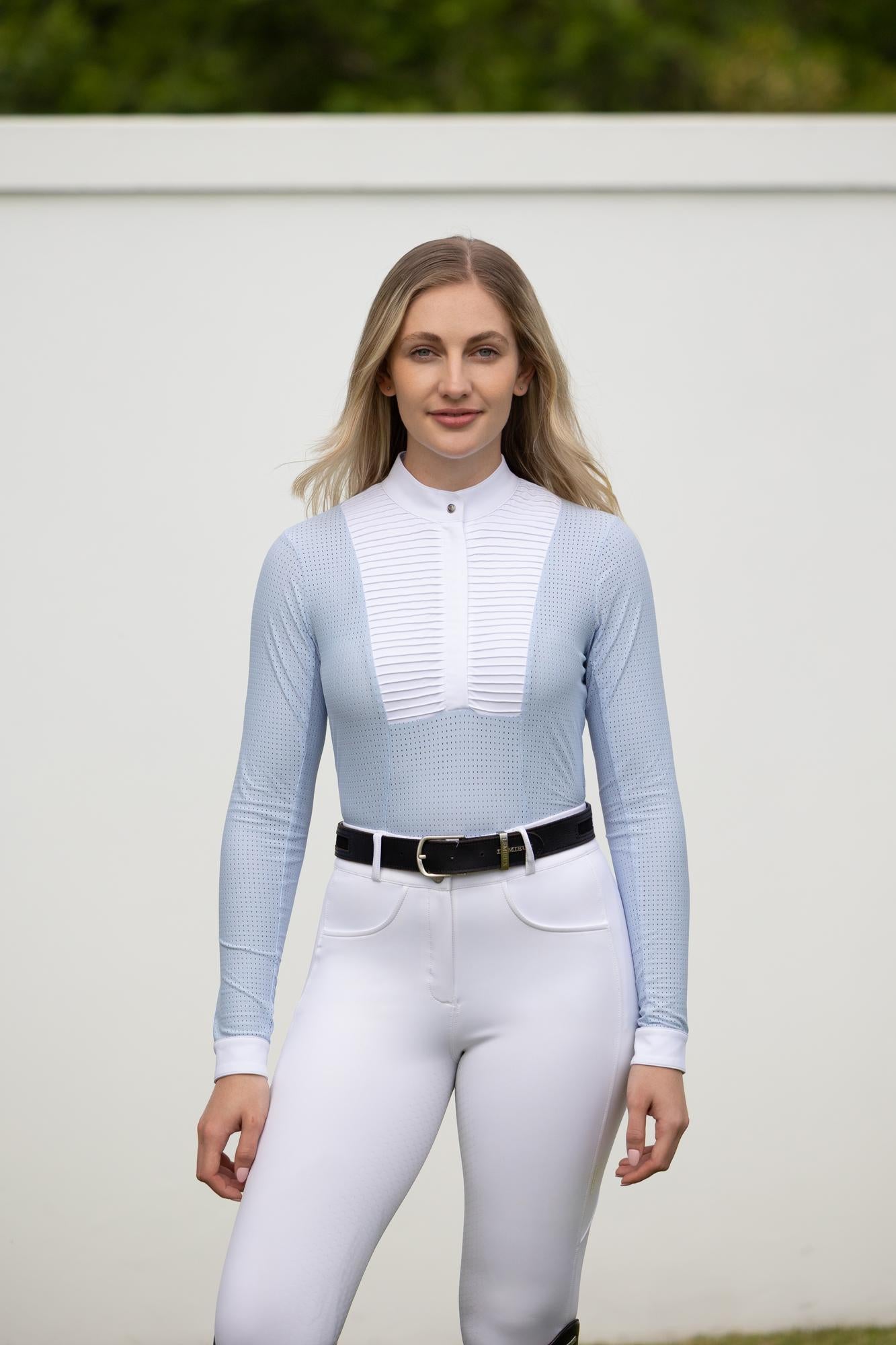 Model wearing LeMieux Arielle Long Sleeve Show Shirt in Mist with white stand collar and pleated bib front, paired with white breeches and black belt