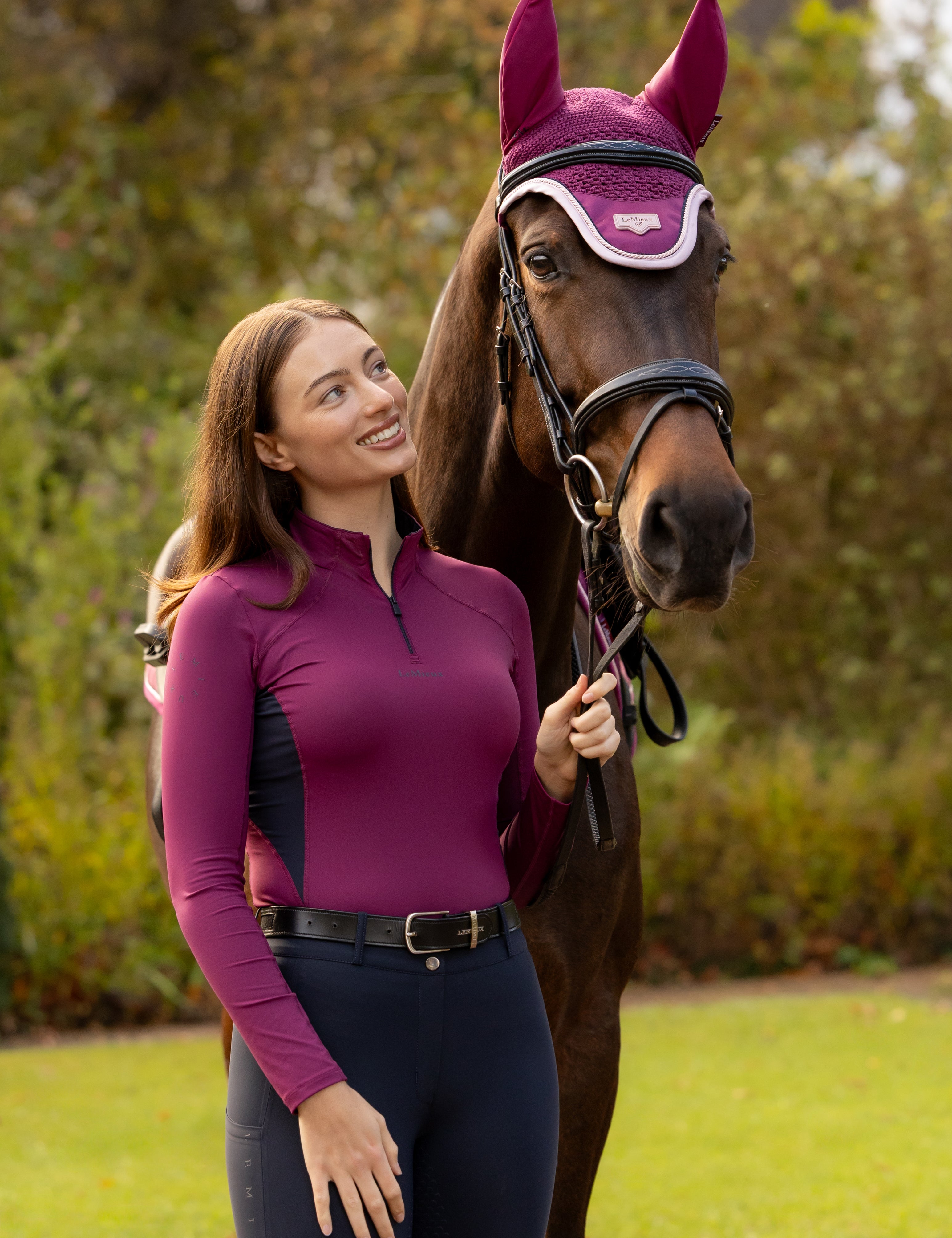 Front view of a female model wearing the Blackberry LeMieux Classique Base Layer while standing next to a horse outside