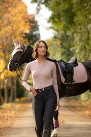 Front view of a female model wearing the Wild Rose LeMieux Lisa Long Sleeve Base Layer while walking away from a black horse on a path outside