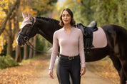 Front view of a female model wearing the Wild Rose LeMieux Lisa Long Sleeve Base Layer while walking away from a black horse on a path outside