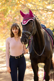 Front view of a female model wearing the Wild Rose LeMieux Lisa Long Sleeve Base Layer while standing next to a black horse outside