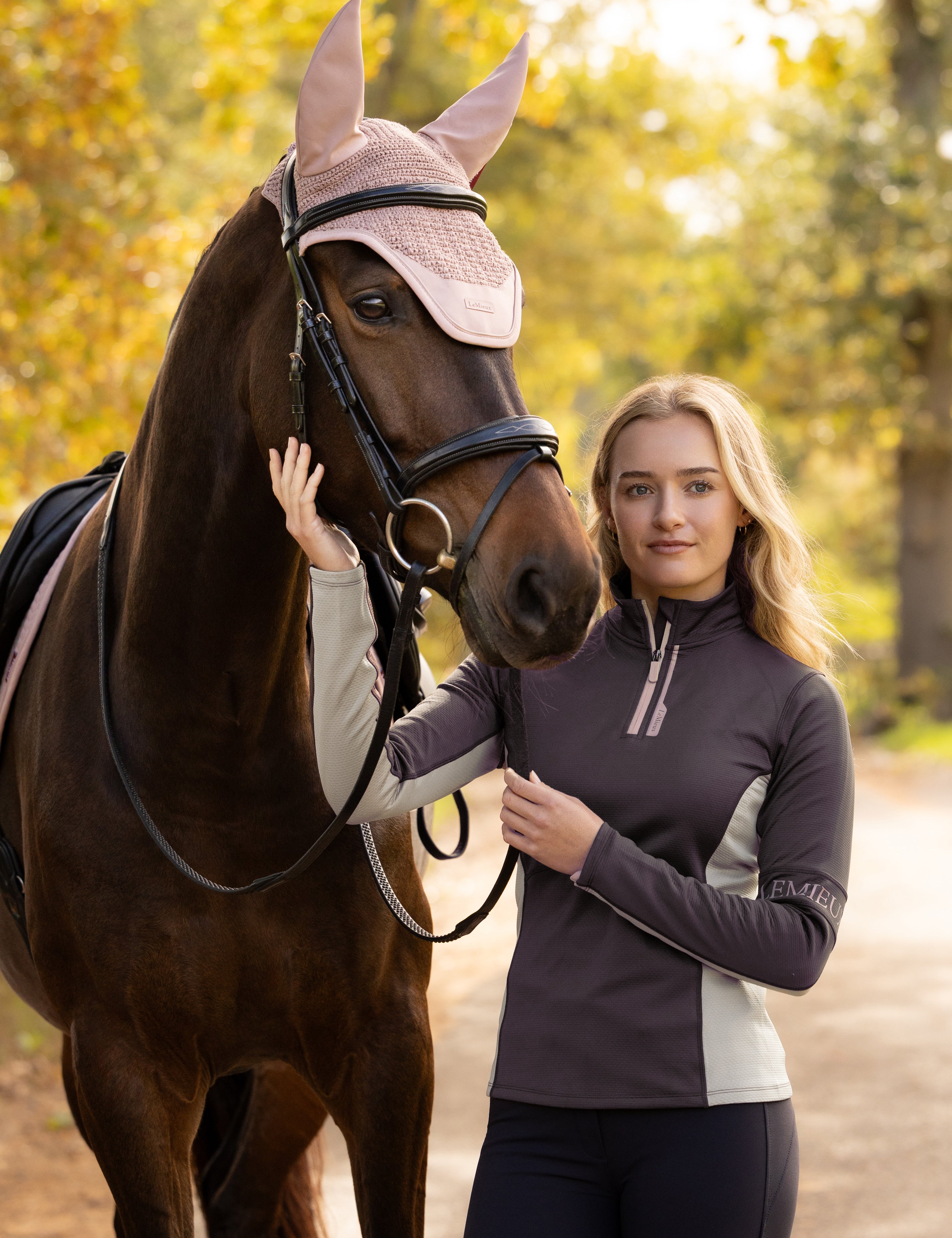 Front view of a female model wearing the Cocoa LeMieux Matilda Mid Layer while standing next to a horse outside