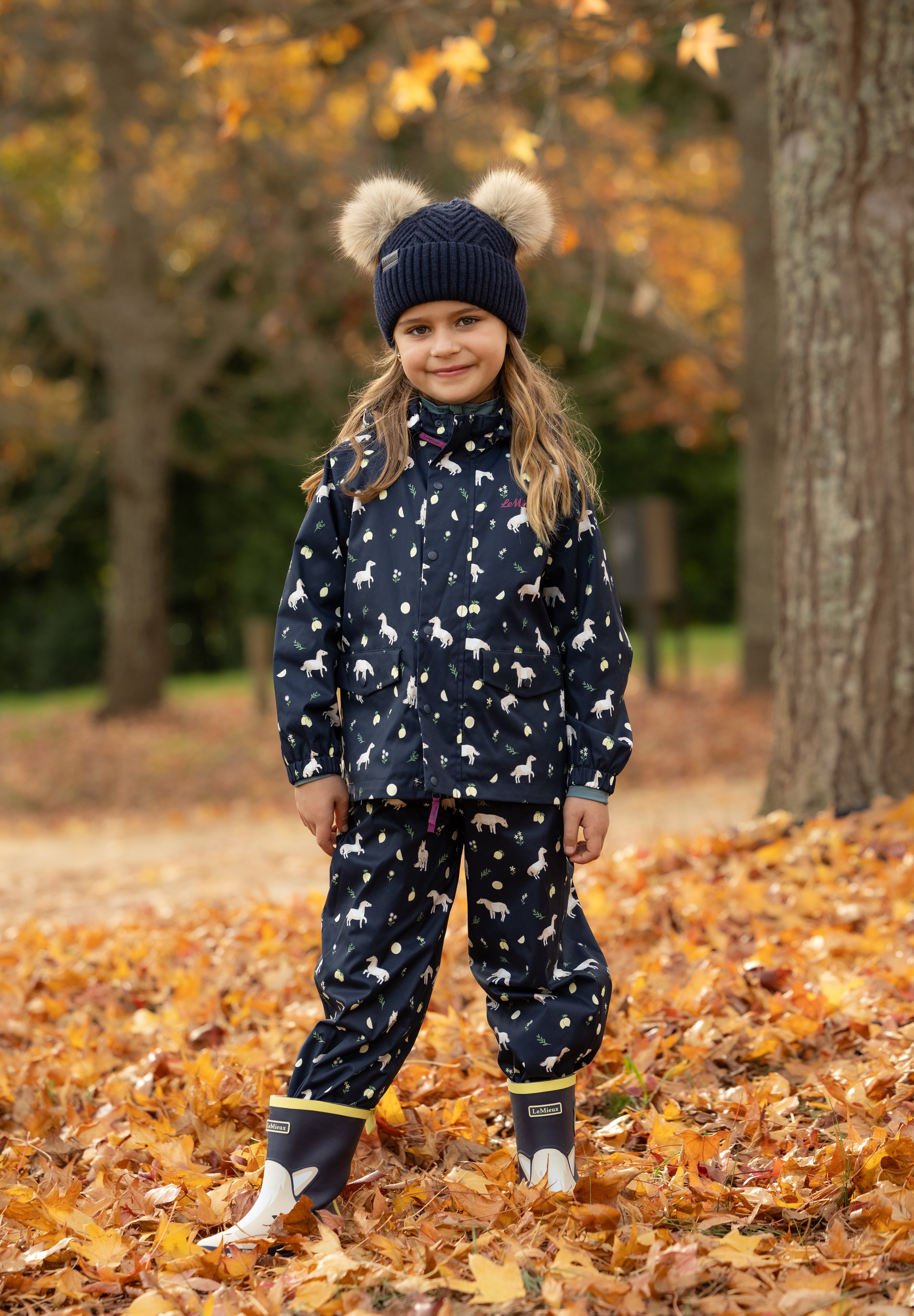 Side view of a young female model wearing Navy LeMieux Mini Pippin Splash Dungarees and standing outdoors