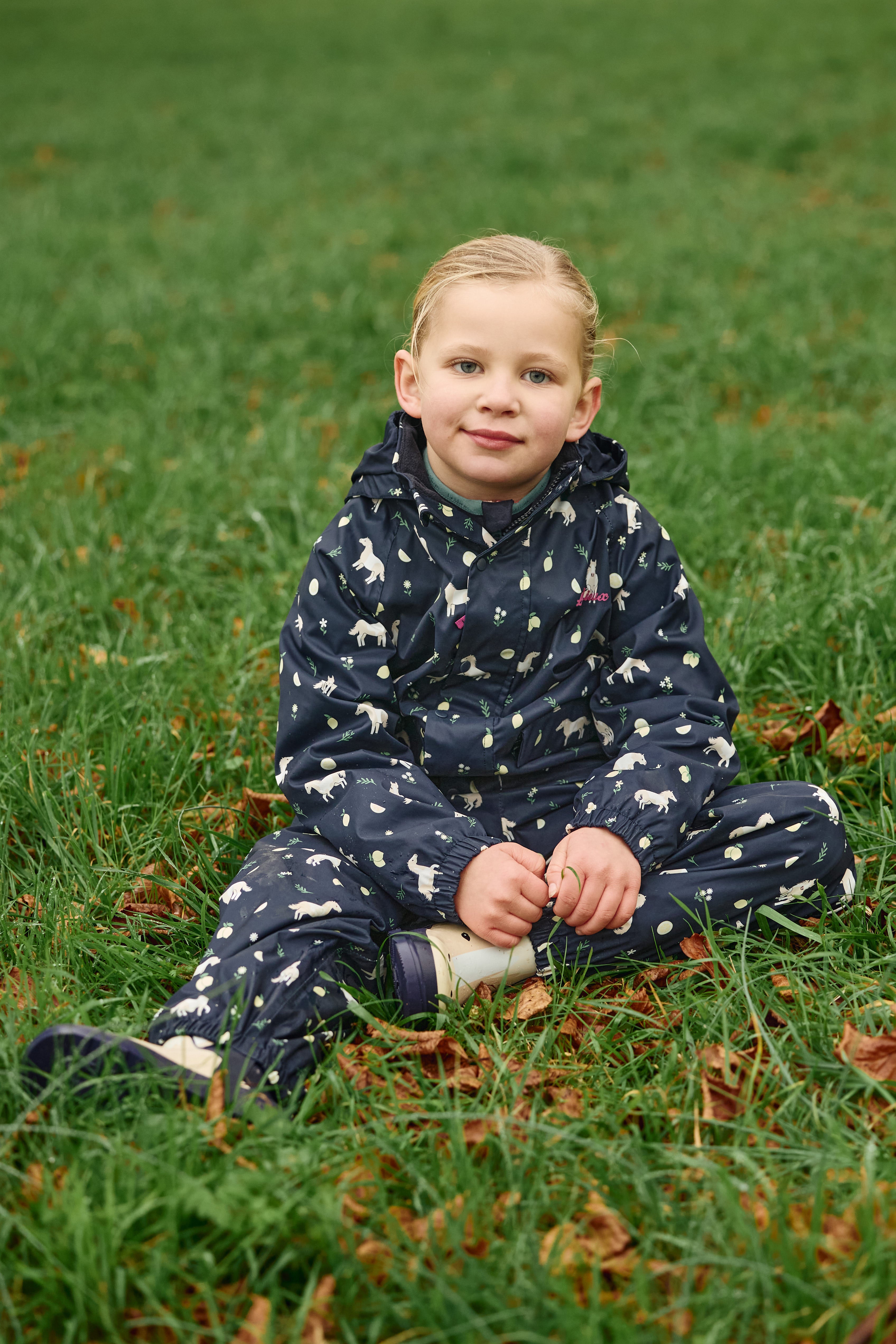 Front view of a young female model wearing Navy LeMieux Mini Pippin Splash Jacket and sitting in the grass