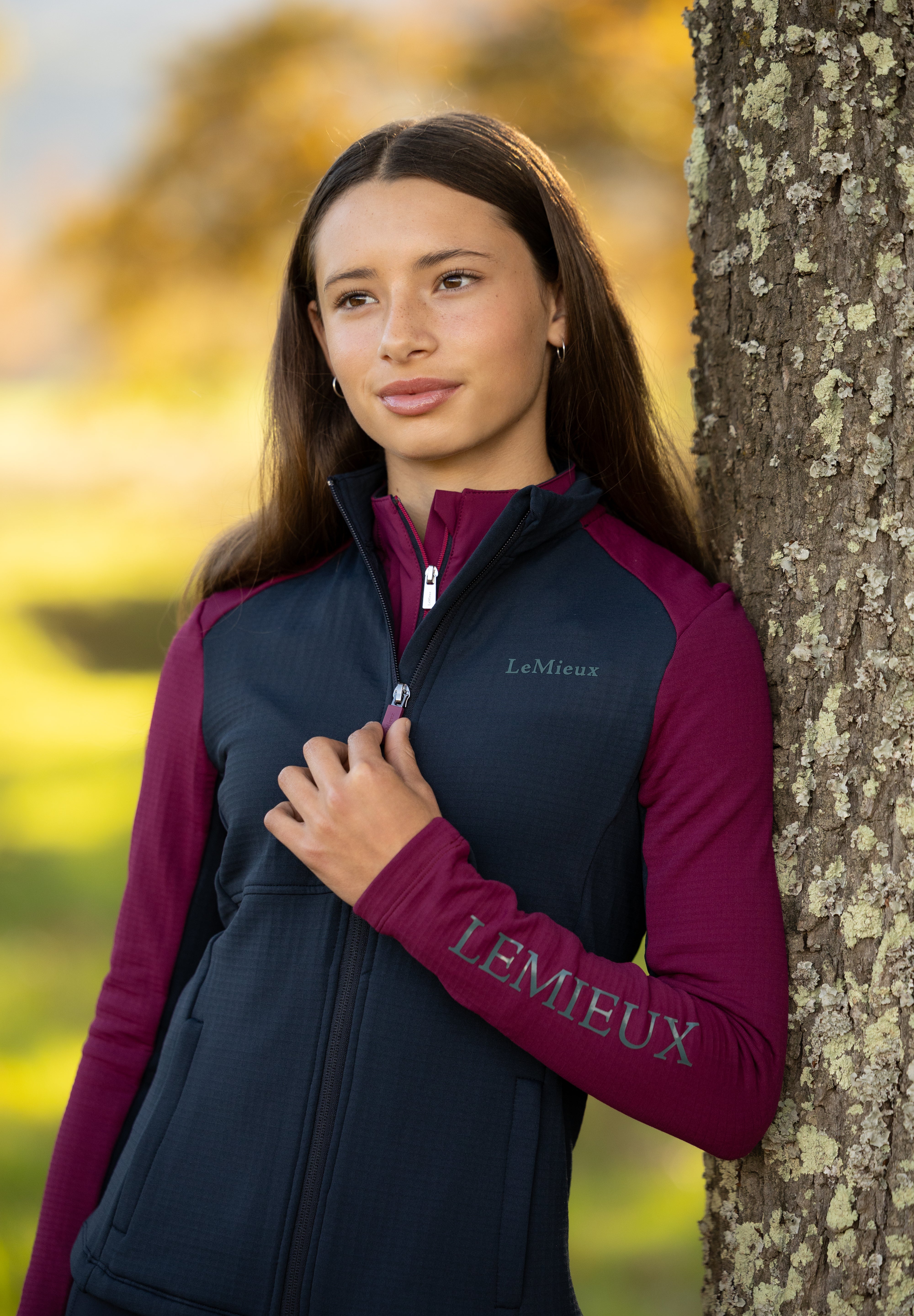 Front view of a model wearing Blackberry LeMieux Young Rider Bethany Fleece Zip Through while leaning against a tree