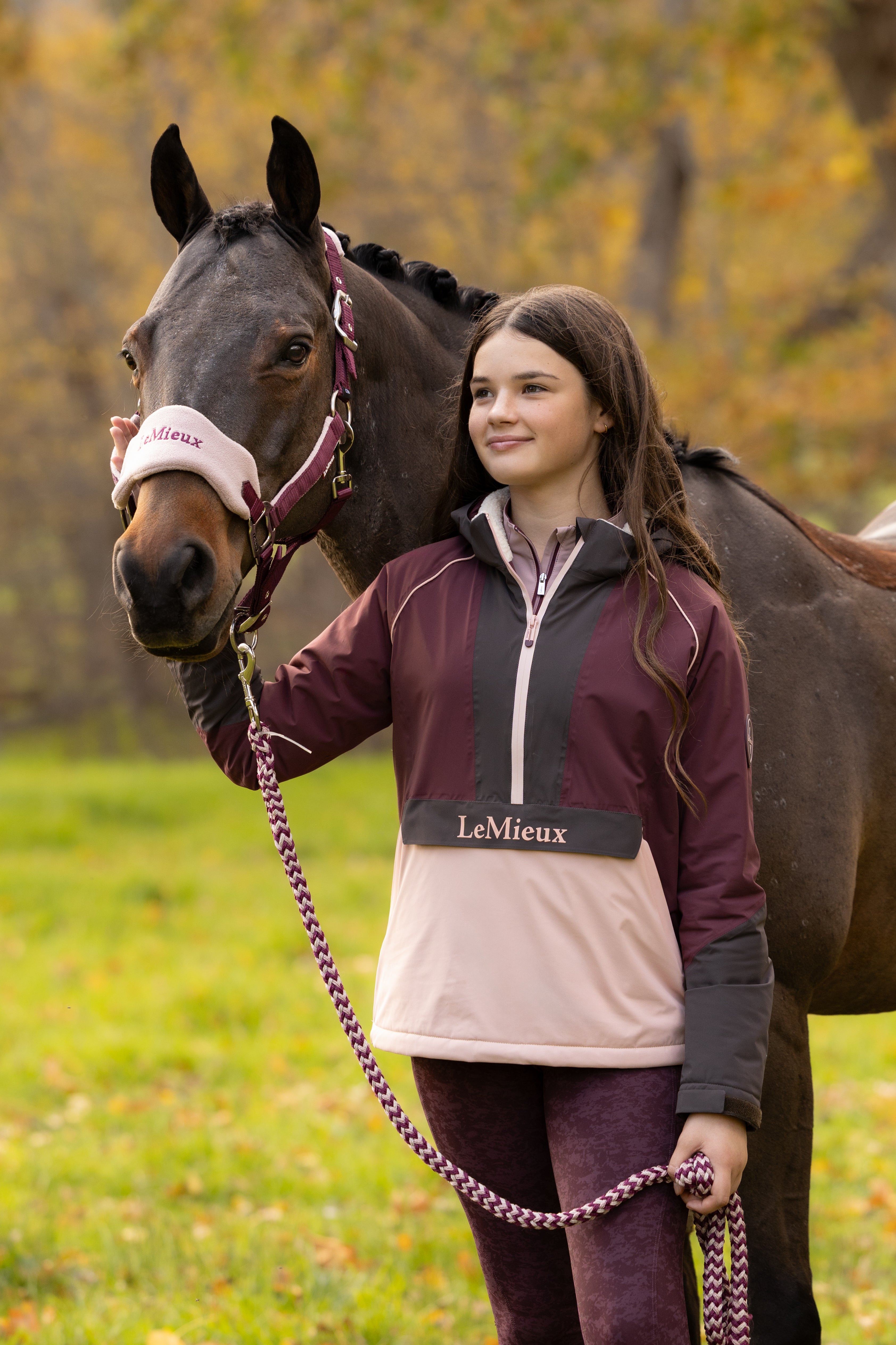 Front view of a young female model wearing Bramble LeMieux Young Rider Bailey Waterproof Anorak next to a horse outdoors