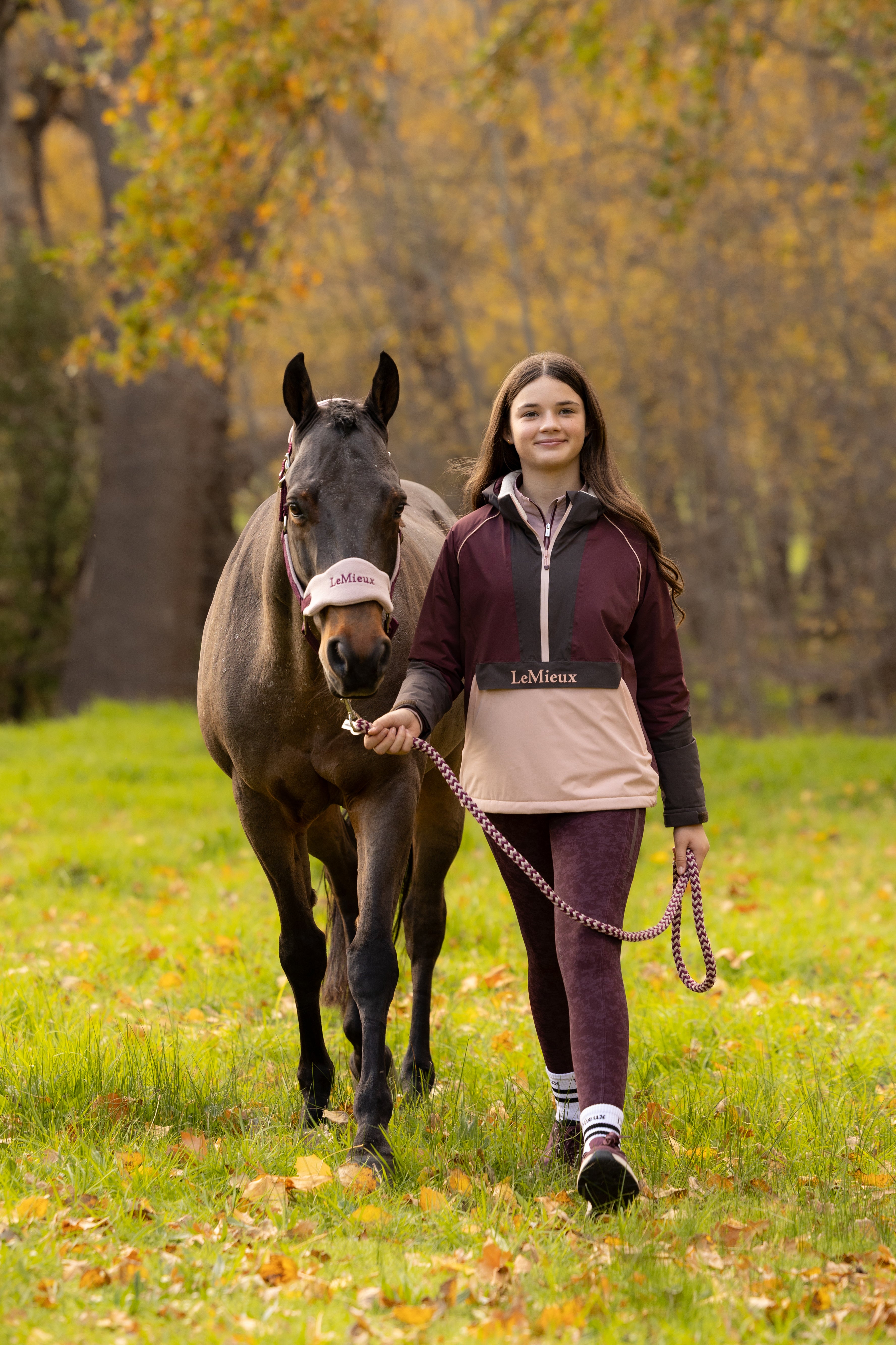 Front view of a young female model wearing Bramble LeMieux Young Rider Bailey Waterproof Anorak next to a horse outdoors