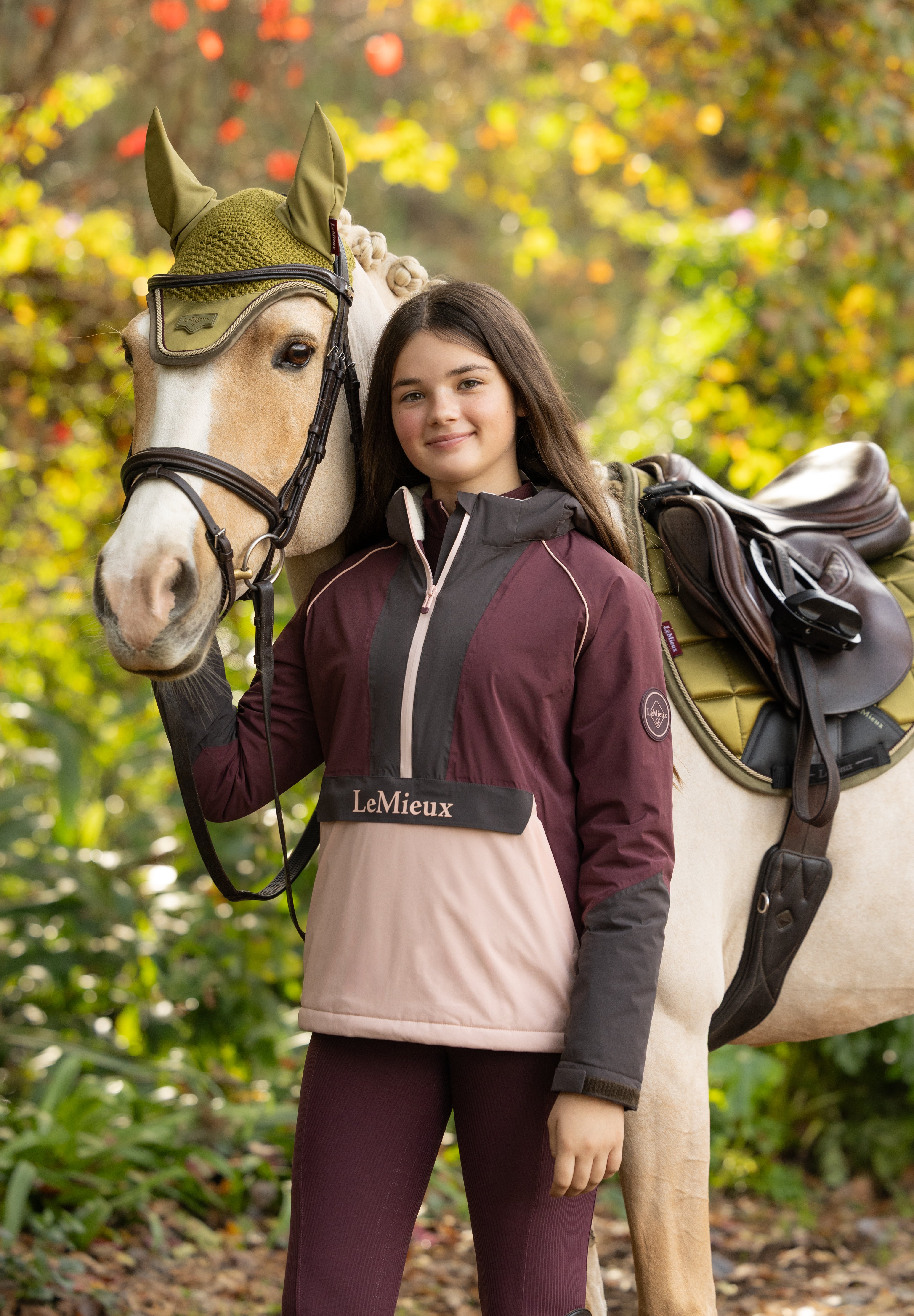 Front view of a young female model wearing Bramble LeMieux Young Rider Bailey Waterproof Anorak next to a horse outdoors