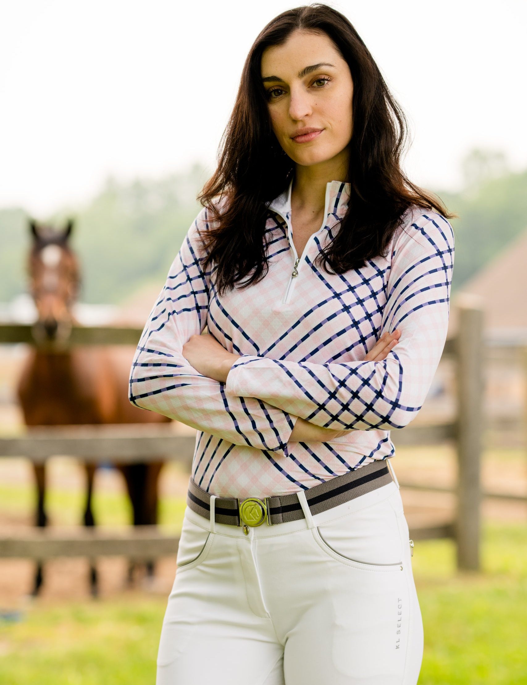 Front view of a female model wearing the white KL Select Gabrielle Breech with a horse standing in the background